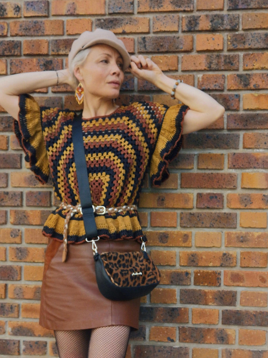 Peut inclure: Une femme porte un béret beige, un haut en crochet marron, jaune et noir, et une jupe en cuir marron. Un sac bandoulière noir avec un rabat imprimé léopard et une ceinture tressée complètent le look. Le fond est un mur de briques.