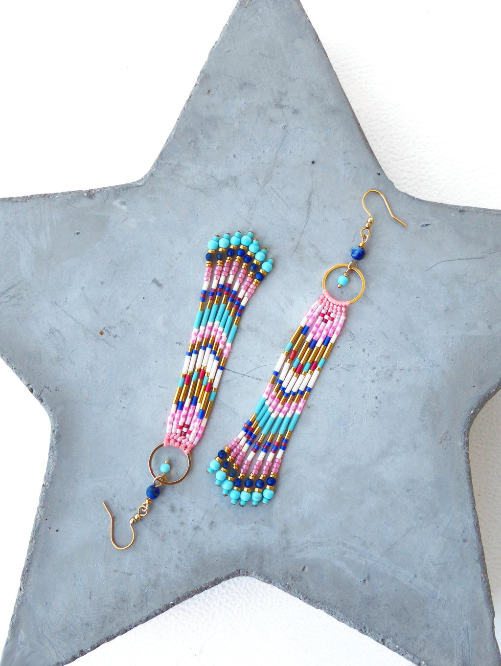 Peut inclure: Une paire de boucles d'oreilles perlées aux couleurs vives. Les boucles d'oreilles présentent des perles roses, turquoise, dorées, blanches et bleu marine. Chaque boucle d'oreille a un anneau doré et une fermeture à crochet. Elles sont posées sur une surface grise en forme d'étoile.