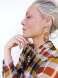 Peut inclure: Une femme porte des boucles d'oreilles à franges perlées avec un arc doré et un accent de perle turquoise. Les boucles d'oreilles présentent un motif de perles orange, jaune, bleu et blanc. Elle porte une chemise à carreaux.