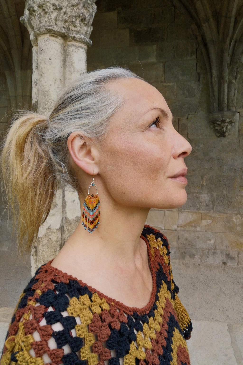 Peut inclure: Une femme portant des boucles d'oreilles en perles en forme de goutte d'eau avec un design à franges colorées. Les boucles d'oreilles présentent des perles orange, jaunes, bleues et marron. Elle porte également un haut en crochet avec un motif géométrique.