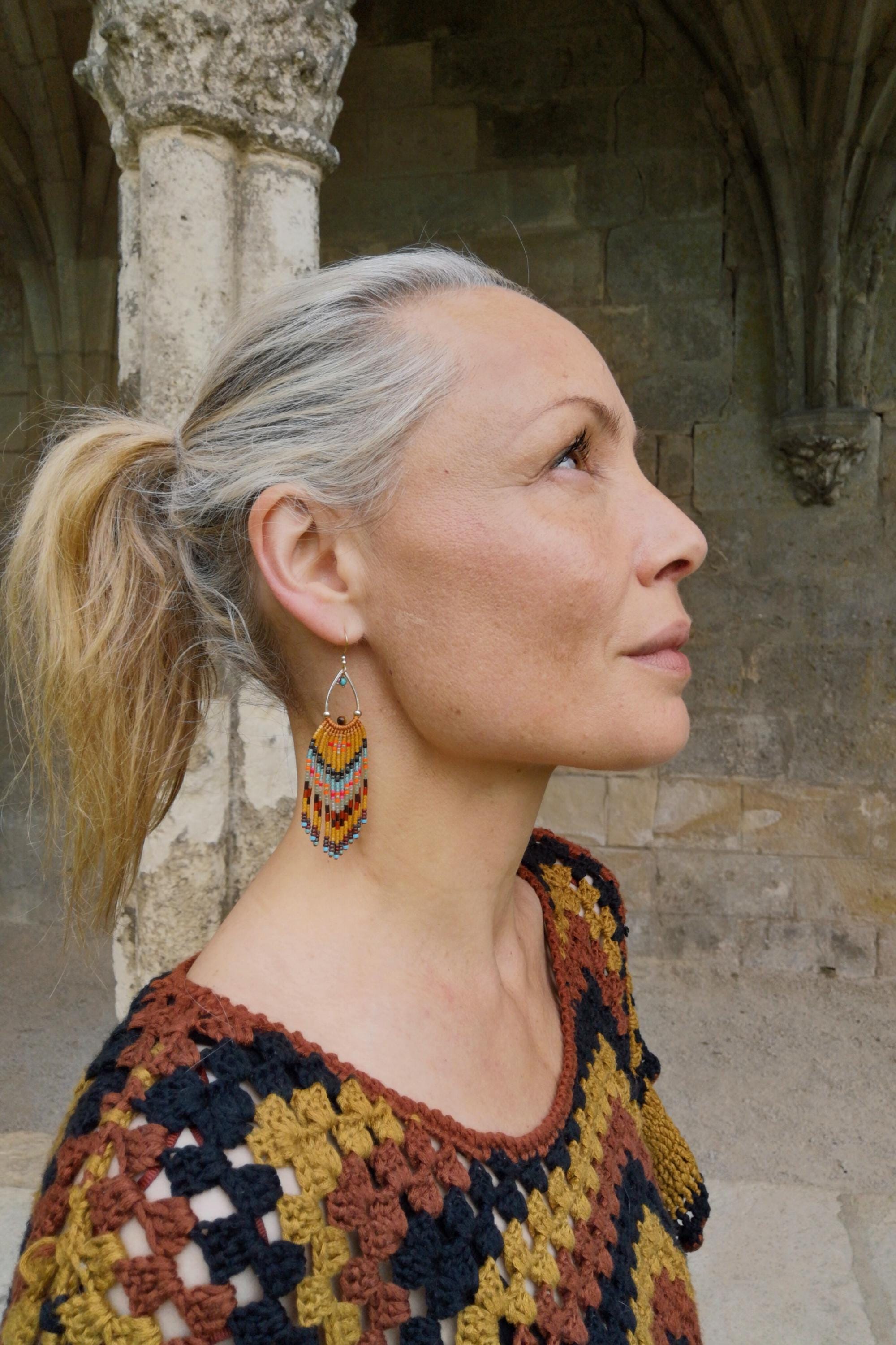 Peut inclure: Une femme portant des boucles d'oreilles en perles en forme de goutte d'eau avec un design à franges colorées. Les boucles d'oreilles présentent des perles orange, jaunes, bleues et marron. Elle porte également un haut en crochet avec un motif géométrique.