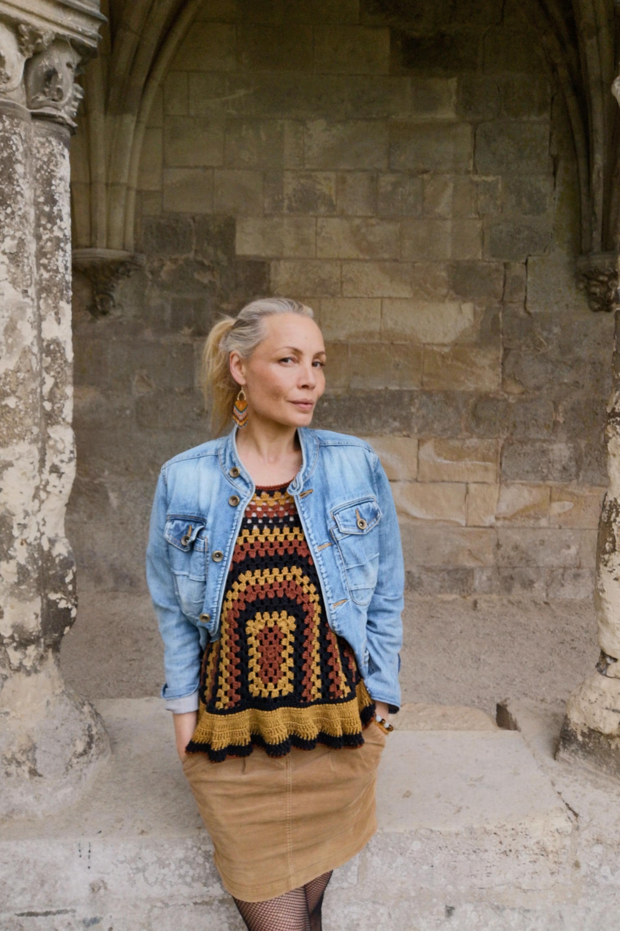 Peut inclure: Une femme porte une veste en jean sur un haut en crochet avec un motif géométrique dans les tons marron, noir et jaune. Elle porte également une jupe beige.