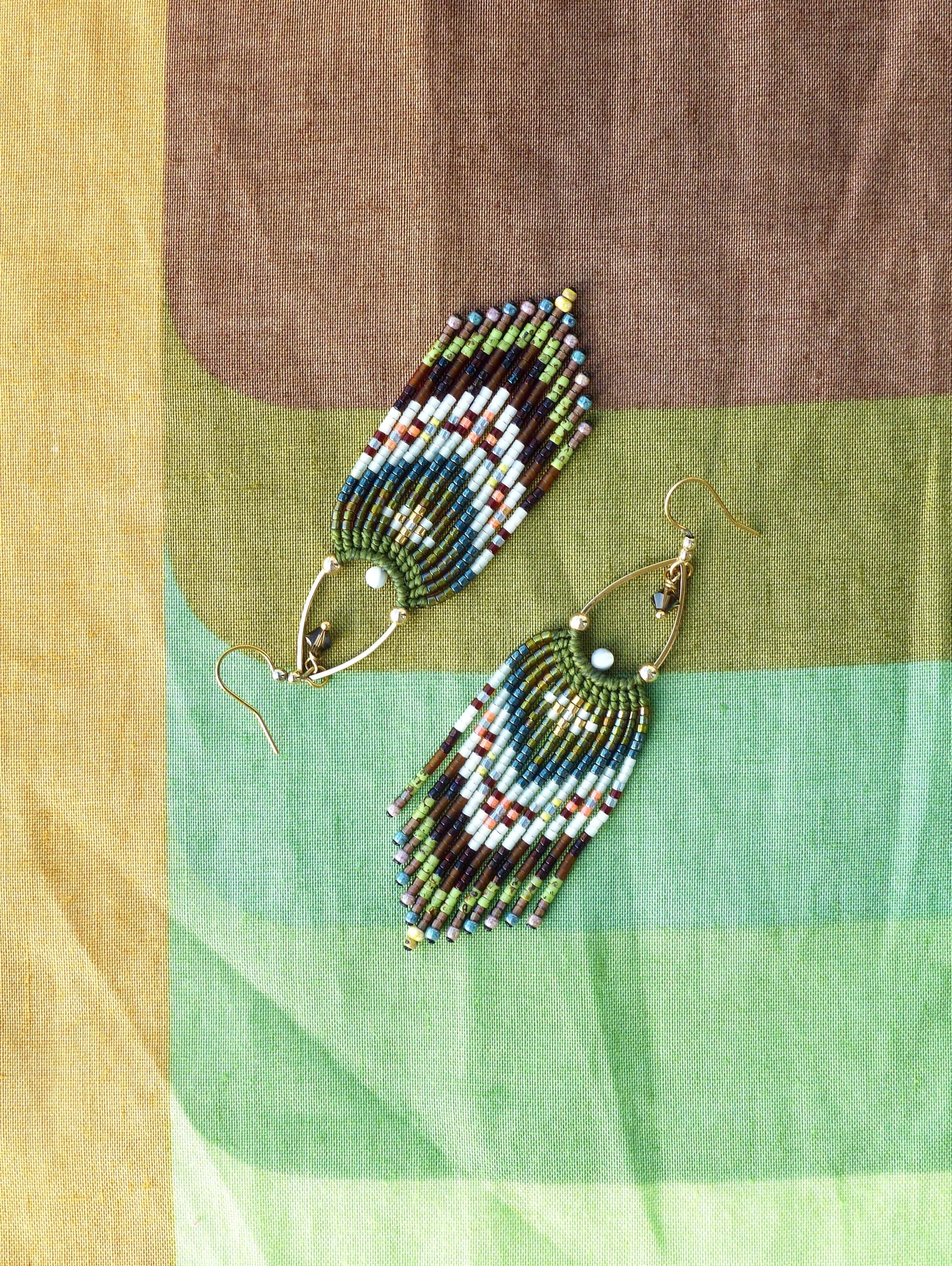 Peut inclure: Une paire de boucles d'oreilles perlées faites à la main. Les boucles d'oreilles ont une forme de goutte d'eau avec un motif de perles blanches, marron, vertes et bleues. Les boucles d'oreilles ont des crochets d'oreilles dorés et sont présentées sur un tissu coloré.