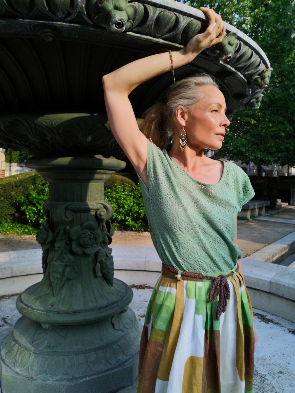 Peut inclure: Une femme porte un haut en dentelle vert clair, une jupe à motifs avec des blocs verts, blancs et marron, et une ceinture tressée marron. Elle porte des boucles d'oreilles dorées et un bracelet. En arrière-plan, une fontaine décorative et des arbres.