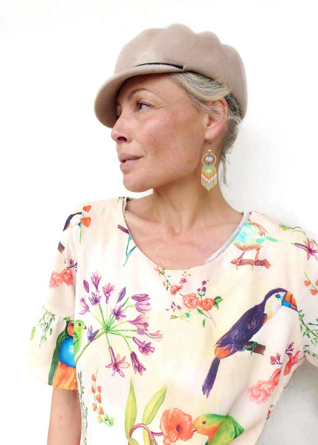 Peut inclure: Une femme porte un béret beige et des boucles d'oreilles en perles colorées. Elle porte un chemisier crème avec un imprimé floral et d'oiseaux. Les boucles d'oreilles ont un cercle doré et des franges colorées.