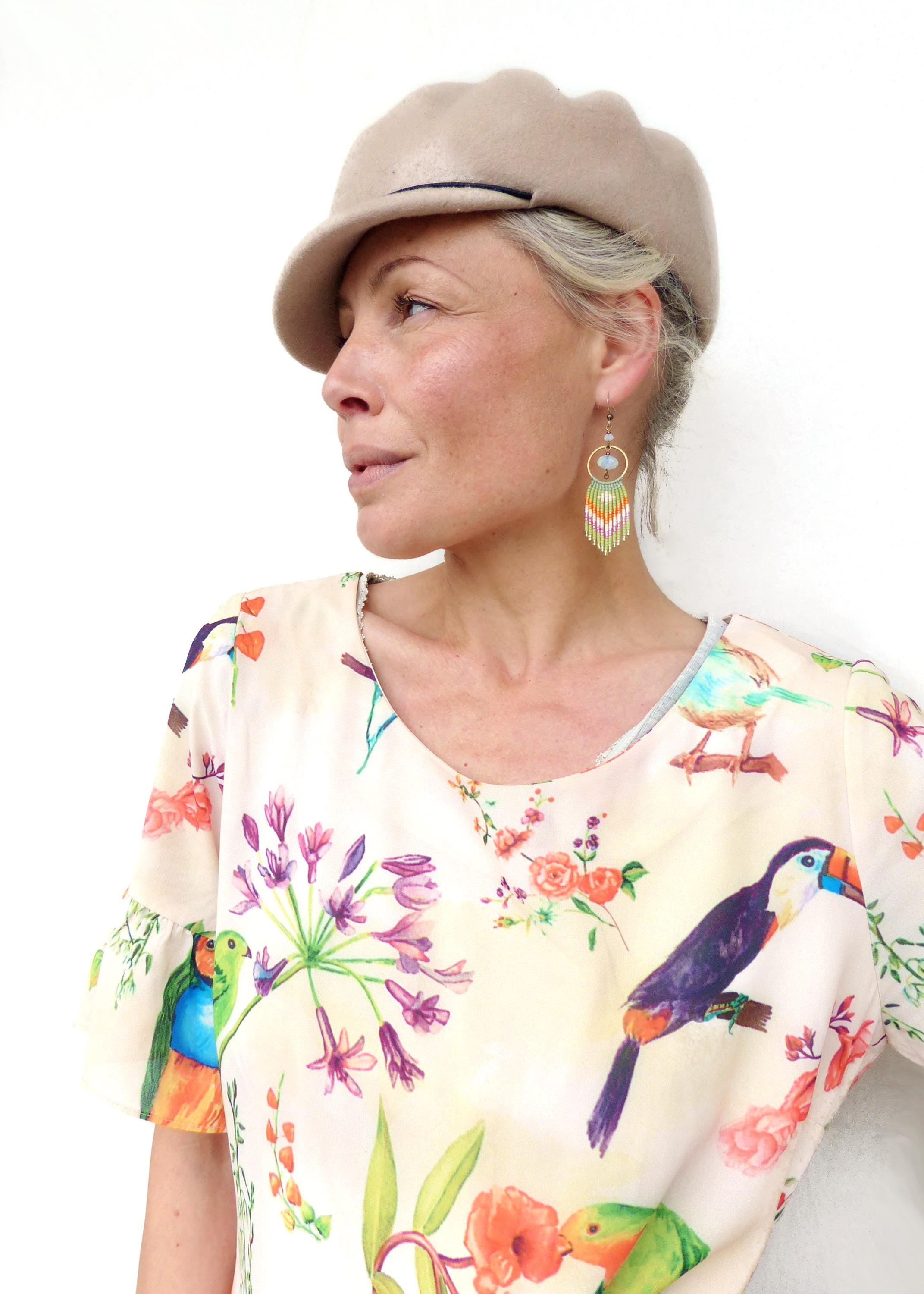 Peut inclure: Une femme porte un béret beige et des boucles d'oreilles en perles colorées. Elle porte un chemisier crème avec un imprimé floral et d'oiseaux. Les boucles d'oreilles ont un cercle doré et des franges colorées.