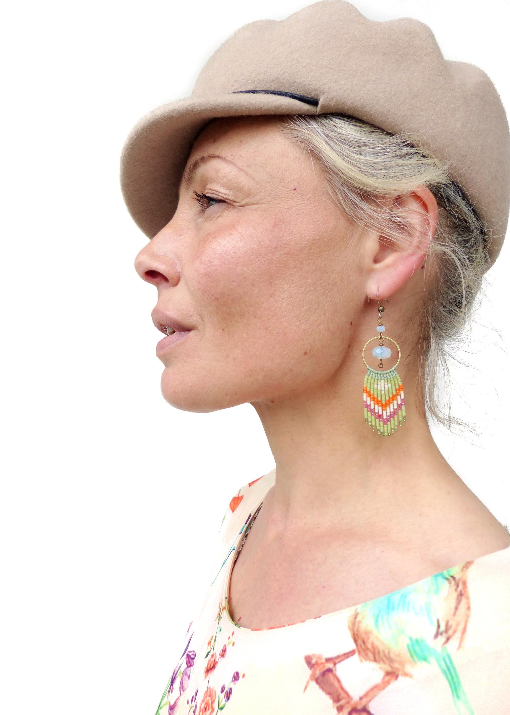 Peut inclure: Une femme porte un chapeau beige et des boucles d'oreilles en perles colorées. Les boucles d'oreilles ont un cercle doré et un motif de perles vertes, oranges, roses et blanches. La femme porte un haut à imprimé floral. Les boucles d'oreilles mesurent environ 7 cm.