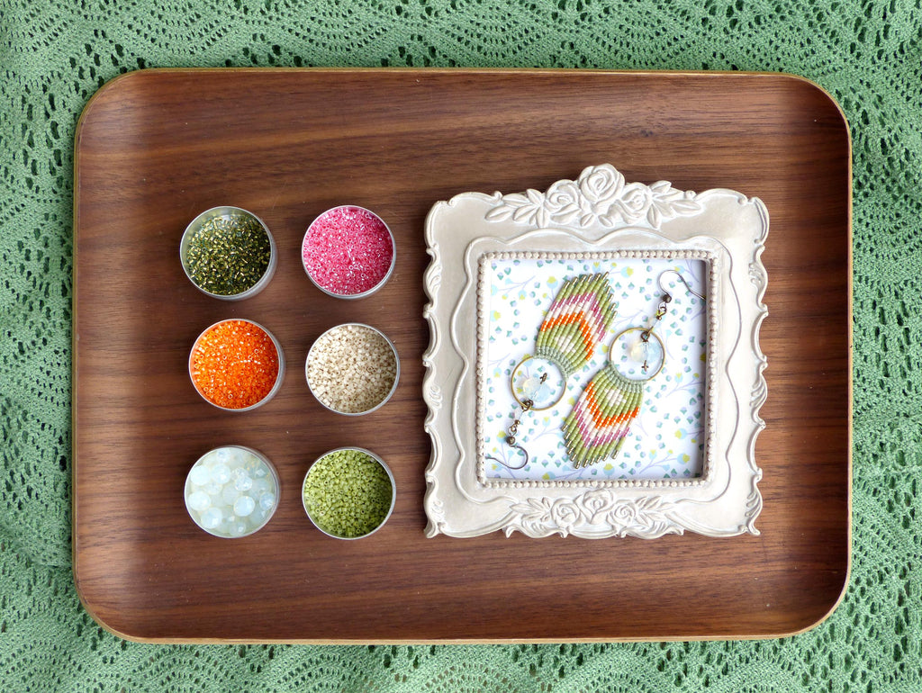Peut inclure: Un plateau en bois présente de petits pots de paillettes colorées et une paire de boucles d'oreilles faites à la main encadrées. Les boucles d'oreilles présentent un motif tissé avec des fils orange, verts et roses. Le fond est un tissu vert à motifs.
