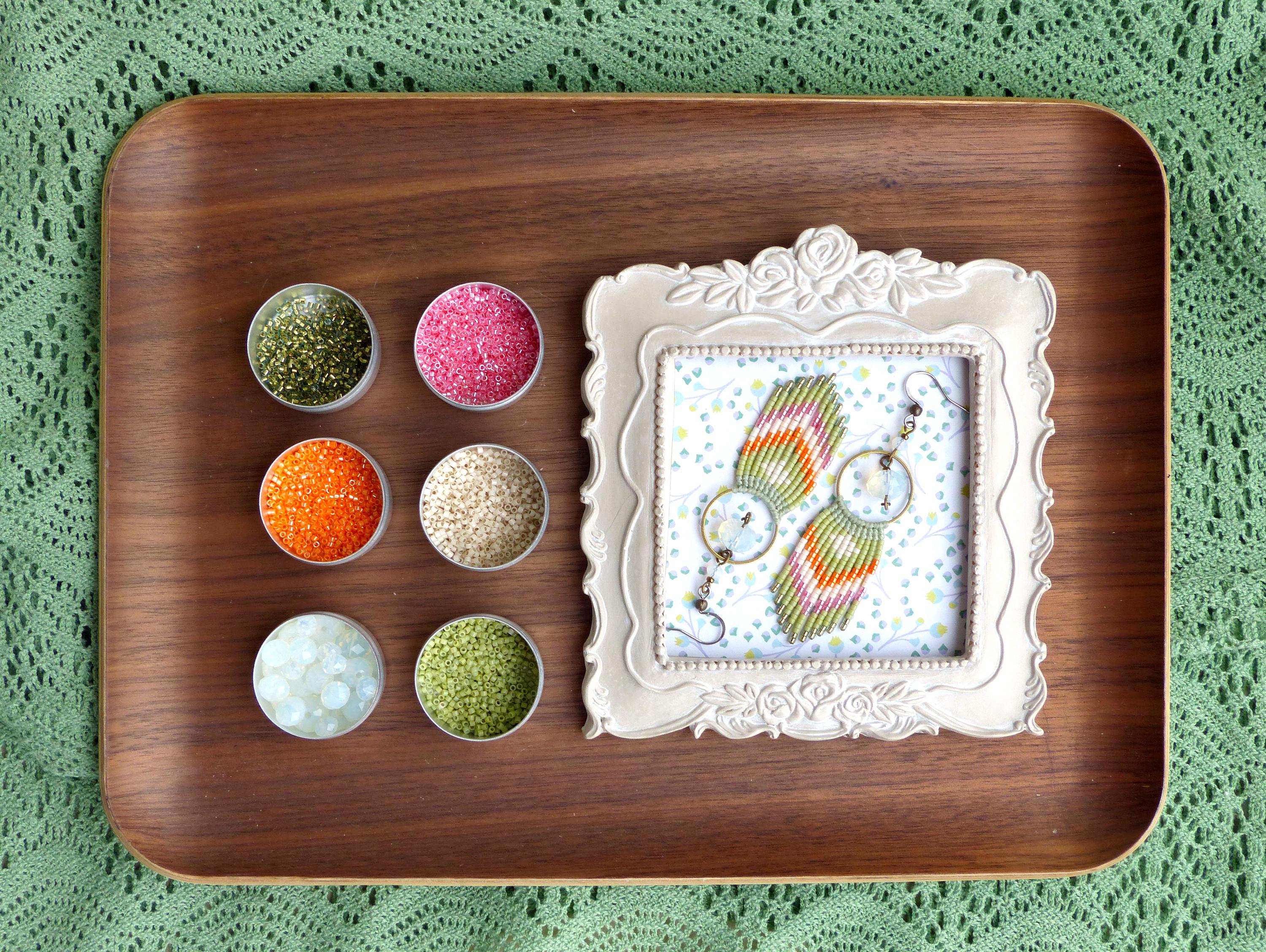 Peut inclure: Un plateau en bois présente de petits pots de paillettes colorées et une paire de boucles d'oreilles faites à la main encadrées. Les boucles d'oreilles présentent un motif tissé avec des fils orange, verts et roses. Le fond est un tissu vert à motifs.
