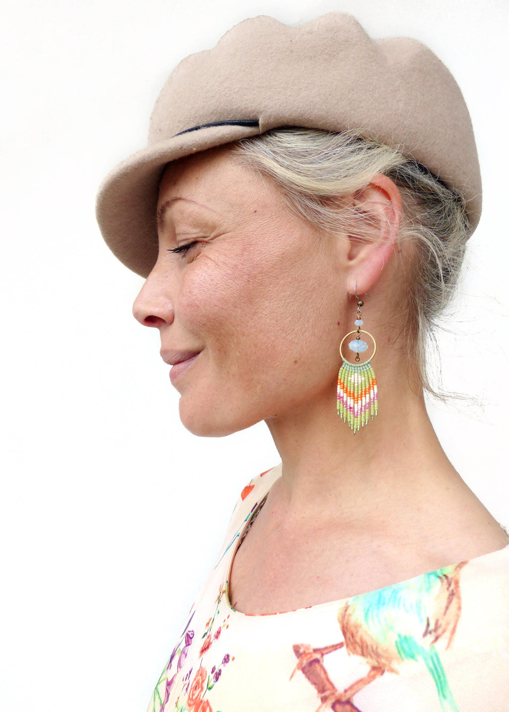 Peut inclure: Une femme porte un chapeau beige et des boucles d'oreilles en perles colorées. Les boucles d'oreilles ont un cercle doré avec une perle bleu clair et une frange de perles vertes, oranges, roses et blanches. Elle porte un haut à motifs floraux.