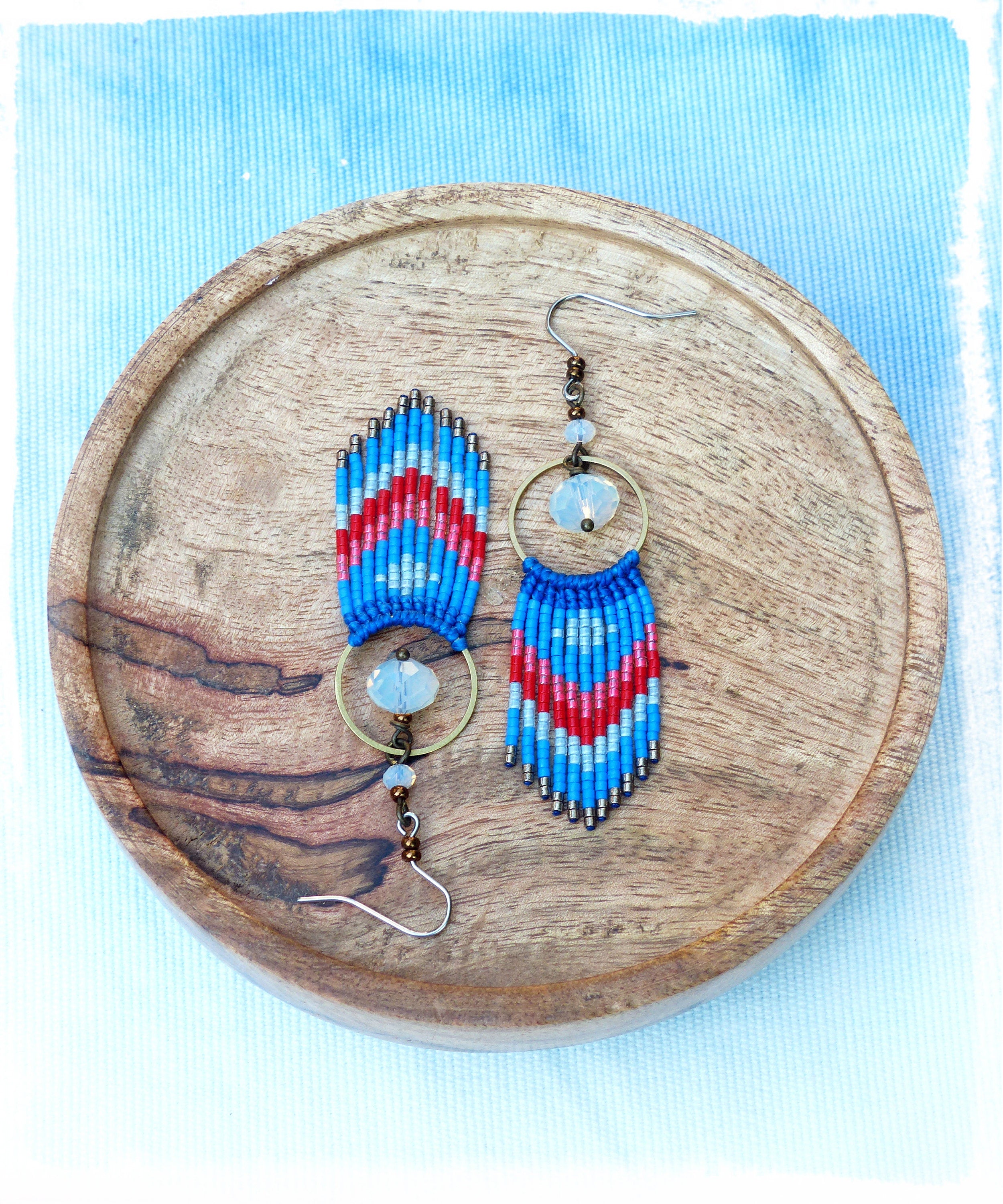 Peut inclure: Une paire de boucles d'oreilles en perles bleues, rouges et blanches avec un charme en cristal transparent suspendu à chaque boucle d'oreille. Les boucles d'oreilles sont présentées sur une surface en bois.