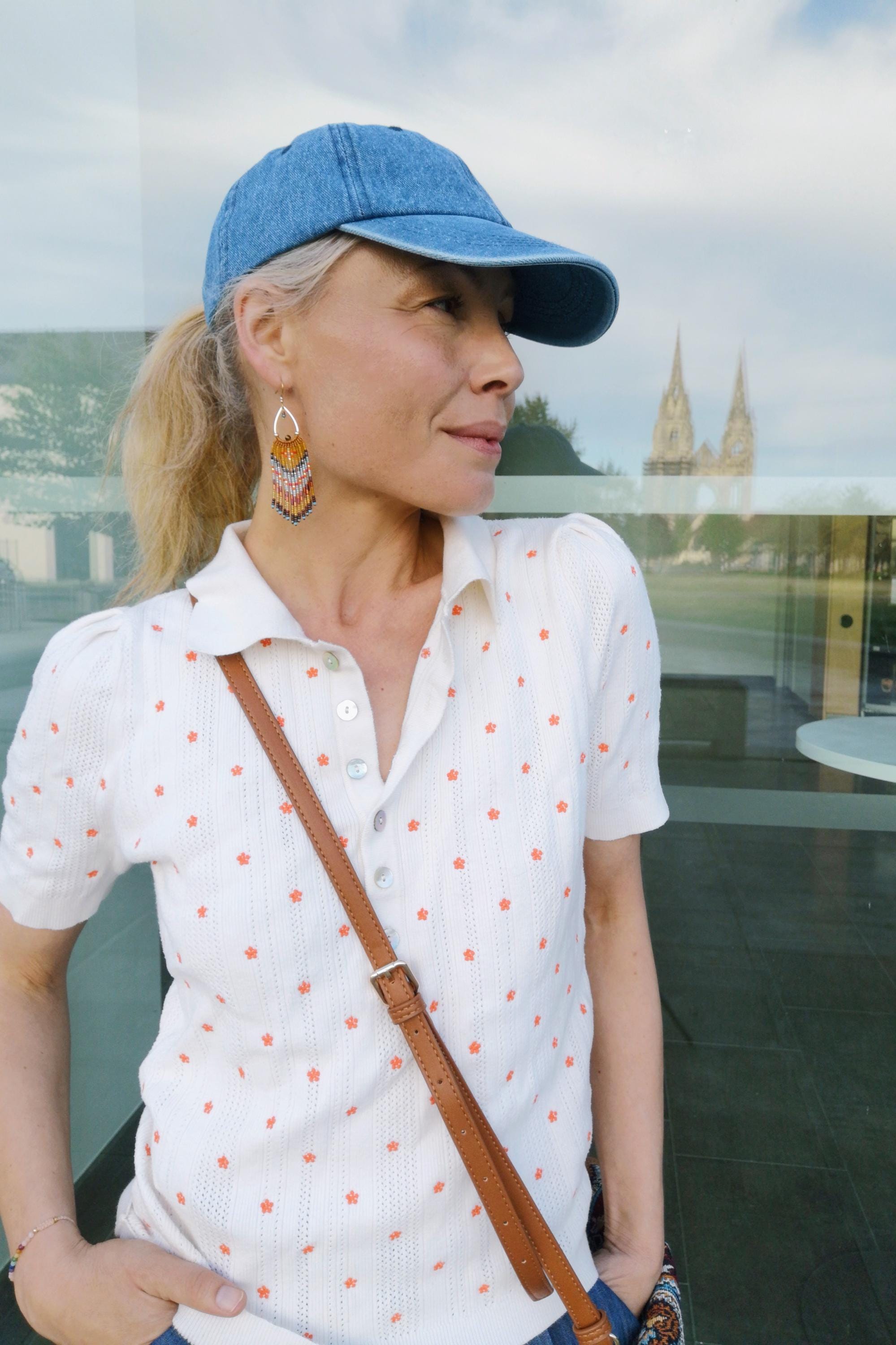 Peut inclure: Casquette de baseball en jean bleu clair et polo blanc à motifs floraux orange. La personne porte des boucles d'oreilles dorées avec des perles colorées et un sac à bandoulière en cuir marron. En arrière-plan, un bâtiment à deux flèches.