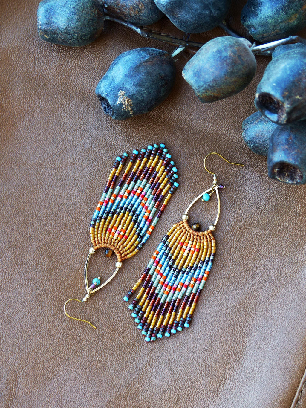 Peut inclure: Une paire de boucles d'oreilles perlées avec des crochets dorés et un design en forme de goutte d'eau. Les boucles d'oreilles présentent une frange de perles colorées dans les tons marron, orange, bleu et turquoise. Elles sont posées sur une surface en cuir marron.