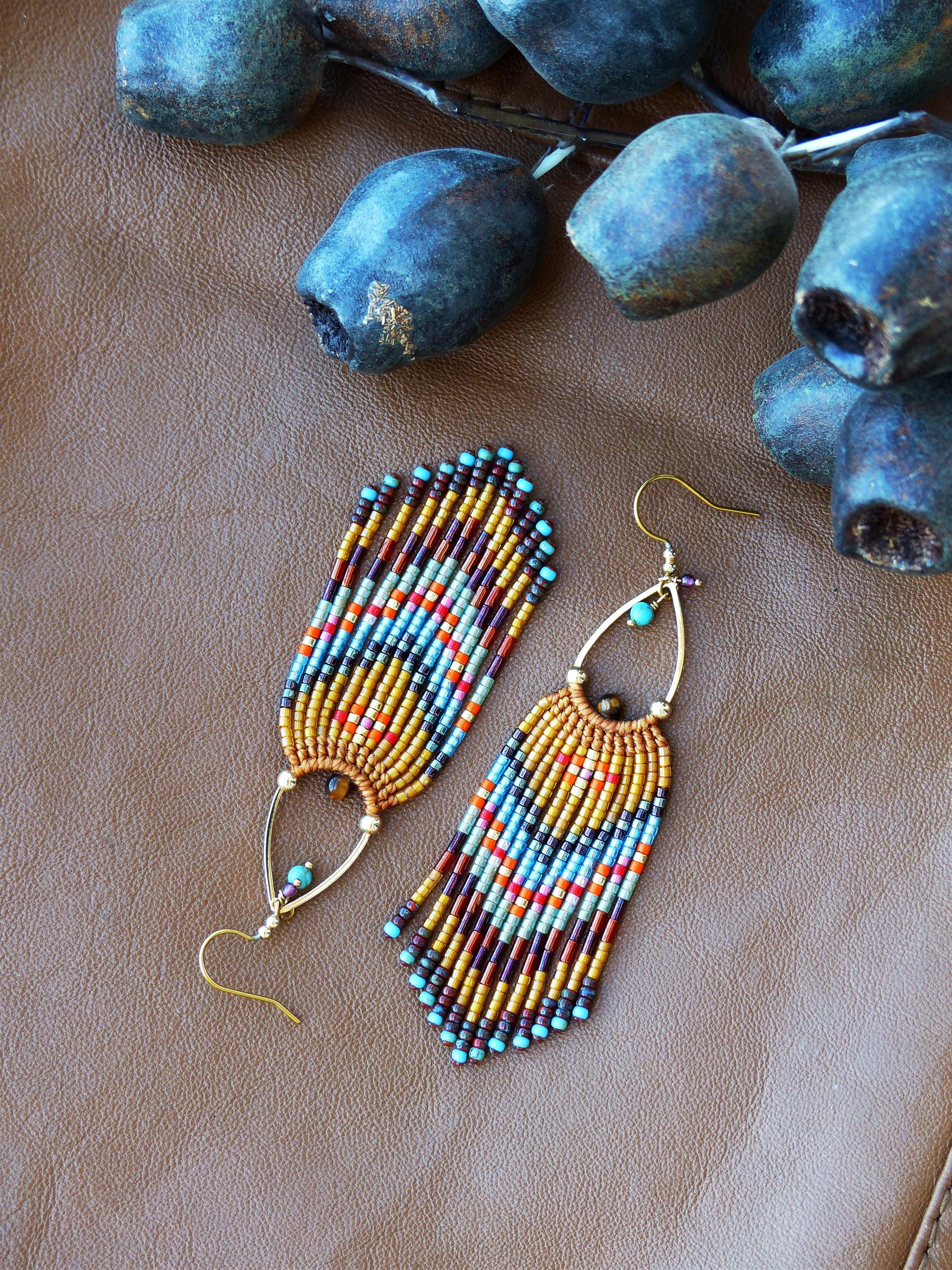 Peut inclure: Une paire de boucles d'oreilles perlées avec des crochets dorés et un design en forme de goutte d'eau. Les boucles d'oreilles présentent une frange de perles colorées dans les tons marron, orange, bleu et turquoise. Elles sont posées sur une surface en cuir marron.