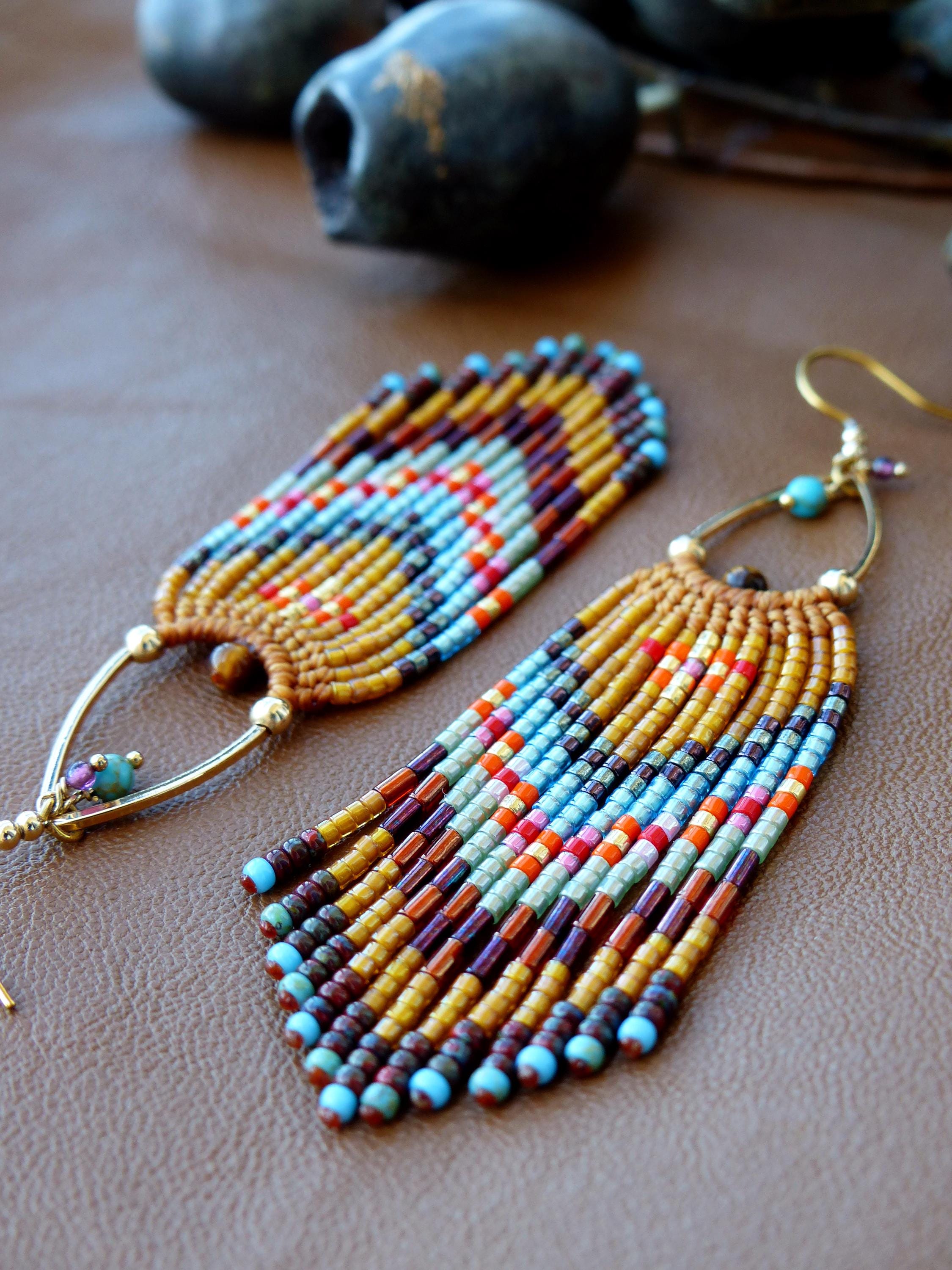 Peut inclure: Gros plan sur deux boucles d'oreilles perlées faites à la main. Les boucles d'oreilles ont des crochets dorés et un design à franges avec des perles bleues, oranges, jaunes et marron. Elles sont posées sur une surface marron.
