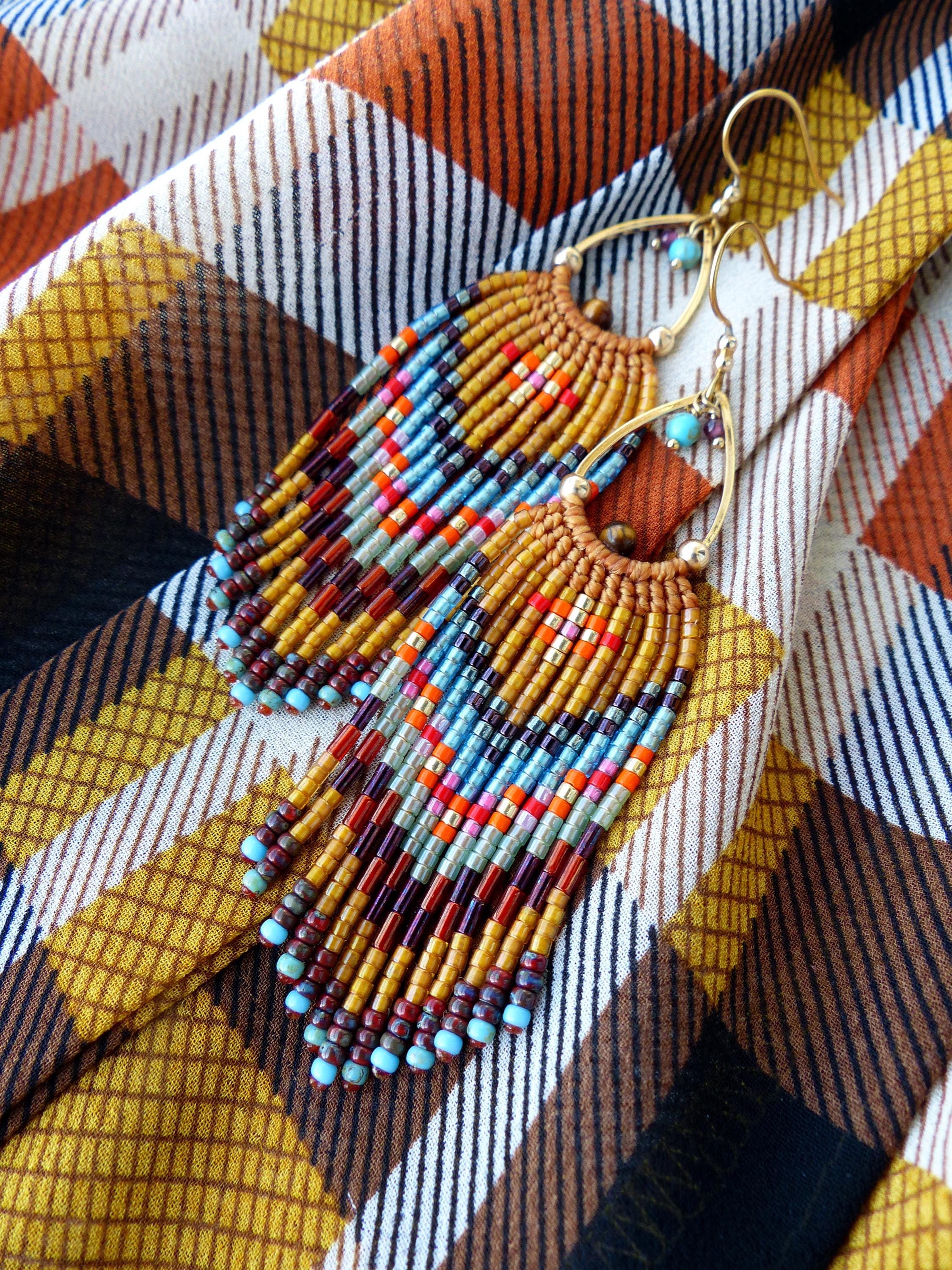 Peut inclure: Boucles d'oreilles en perles faites à la main avec une monture en métal doré. Les boucles d'oreilles présentent un motif de franges avec un mélange de perles colorées dans les tons orange, bleu, rouge et marron. Les boucles d'oreilles sont présentées sur un tissu à carreaux.
