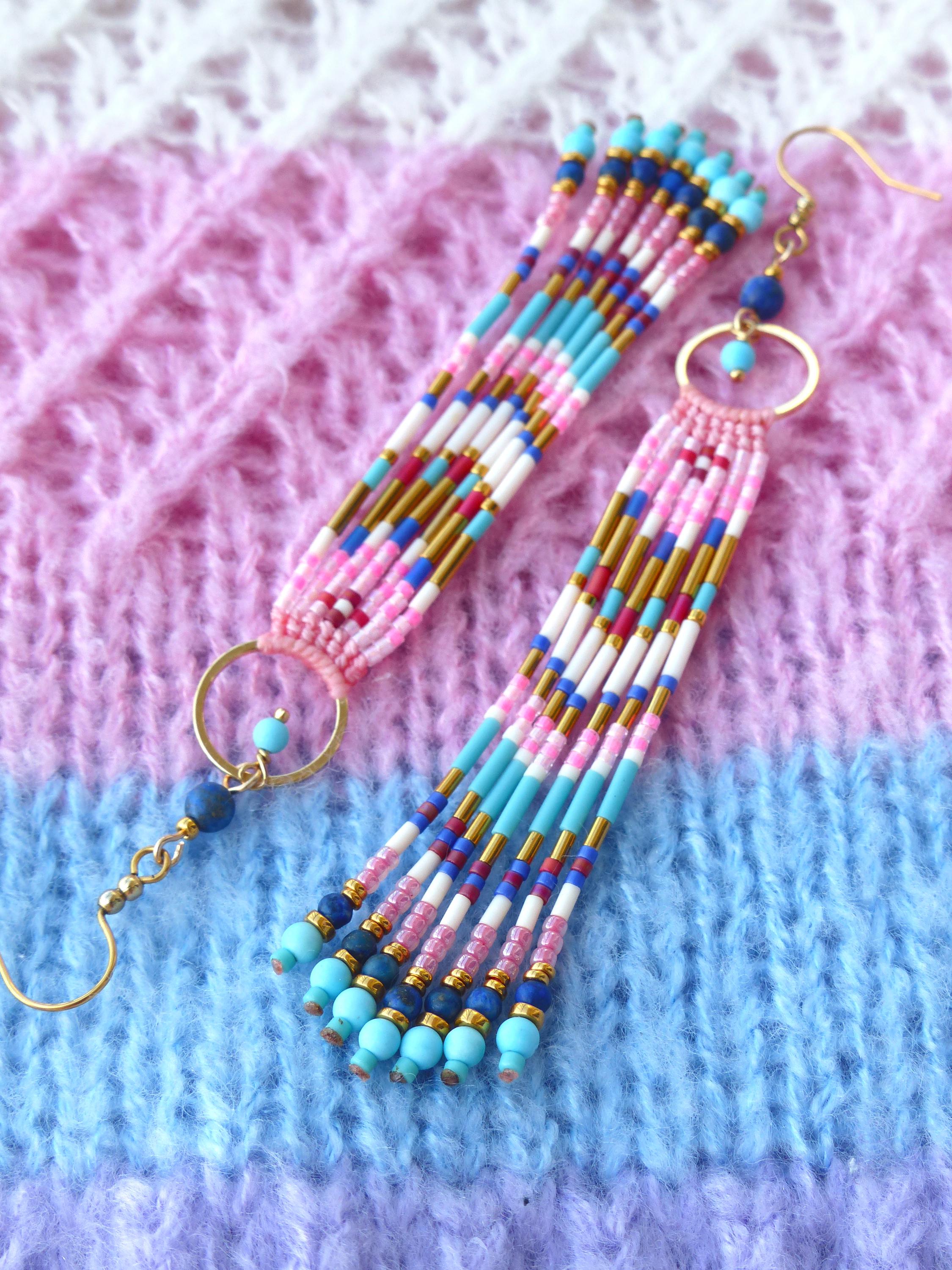 Peut inclure: Une paire de boucles d'oreilles faites à la main. Elles présentent un motif coloré avec des perles roses, bleues, blanches et dorées. Les boucles d'oreilles ont un anneau doré et une breloque perlée. Le fond est un tissu tricoté rose, bleu et violet.