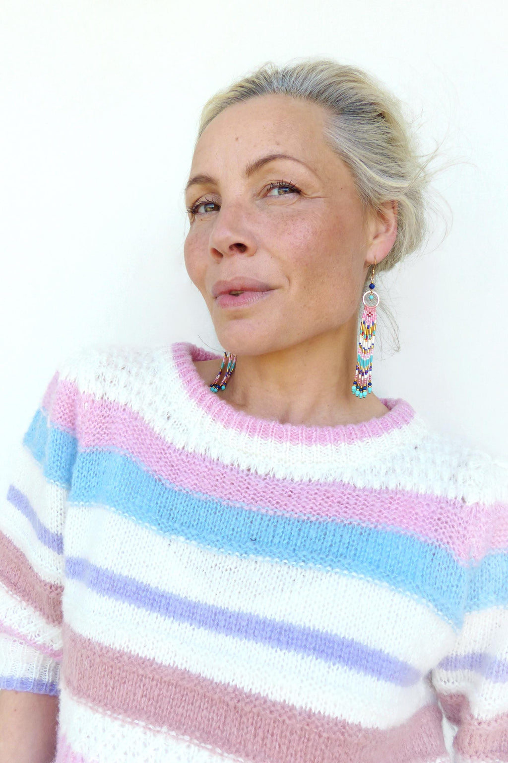Peut inclure: Une femme porte un pull rayé avec des rayures horizontales roses, blanches et bleues. Elle porte également de longues boucles d'oreilles en perles avec des perles roses, bleues et jaunes. Le pull a une encolure ronde.