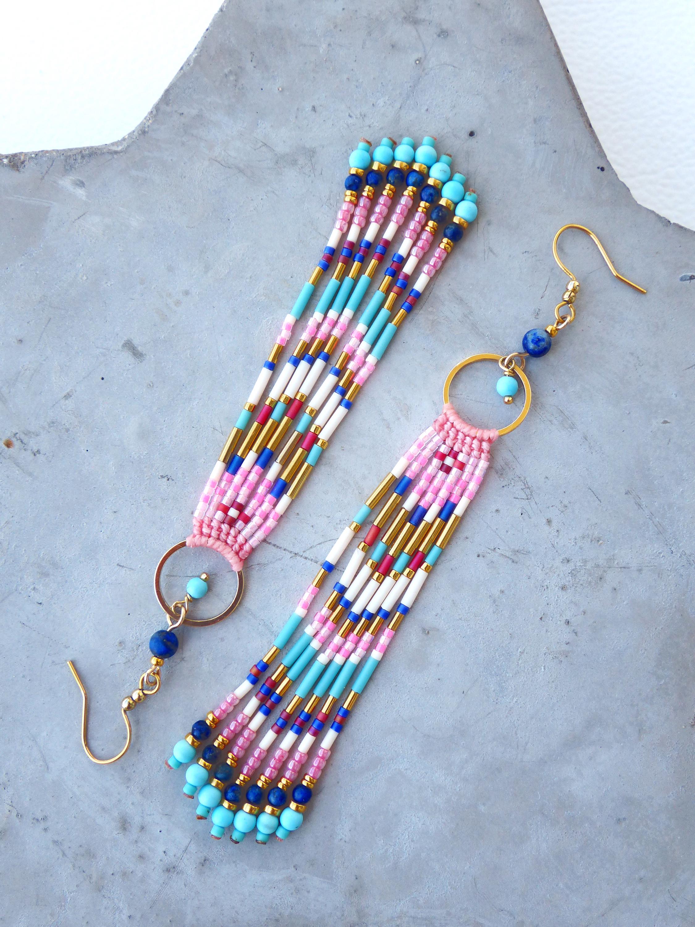 Peut inclure: Une paire de boucles d'oreilles perlées faites à la main. Elles présentent un anneau et un crochet d'oreille dorés, avec des brins de perles roses, bleues, blanches et dorées. Les boucles d'oreilles ont un design à franges et sont ornées de petites perles bleues et dorées.