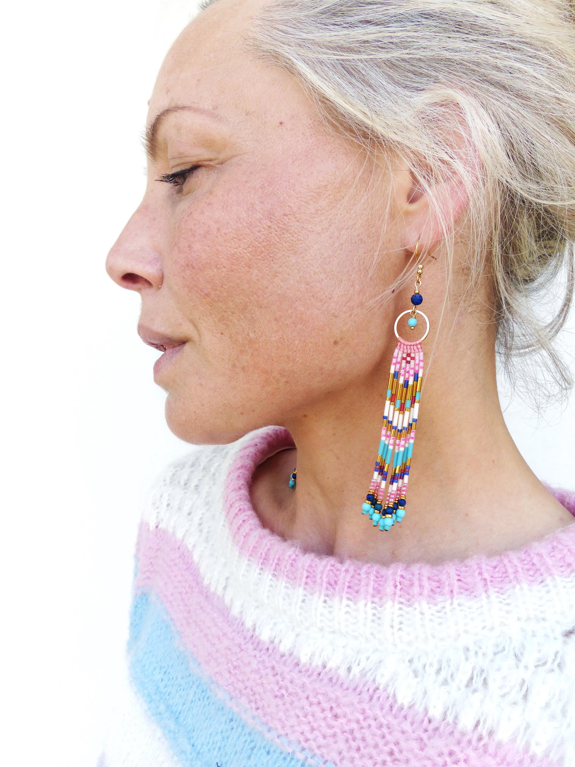 Peut inclure: Longues boucles d'oreilles en perles avec un anneau doré et des franges colorées. Les boucles d'oreilles présentent des perles roses, bleues, turquoise et dorées. La femme porte un pull rayé rose, blanc et bleu.
