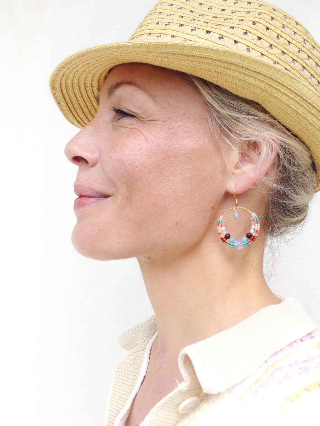 Peut inclure: Profil d'une personne portant un chapeau de paille et des boucles d'oreilles créoles dorées. Les boucles d'oreilles sont ornées de perles colorées rouges, blanches, bleues et marron. La personne porte un pull crème.