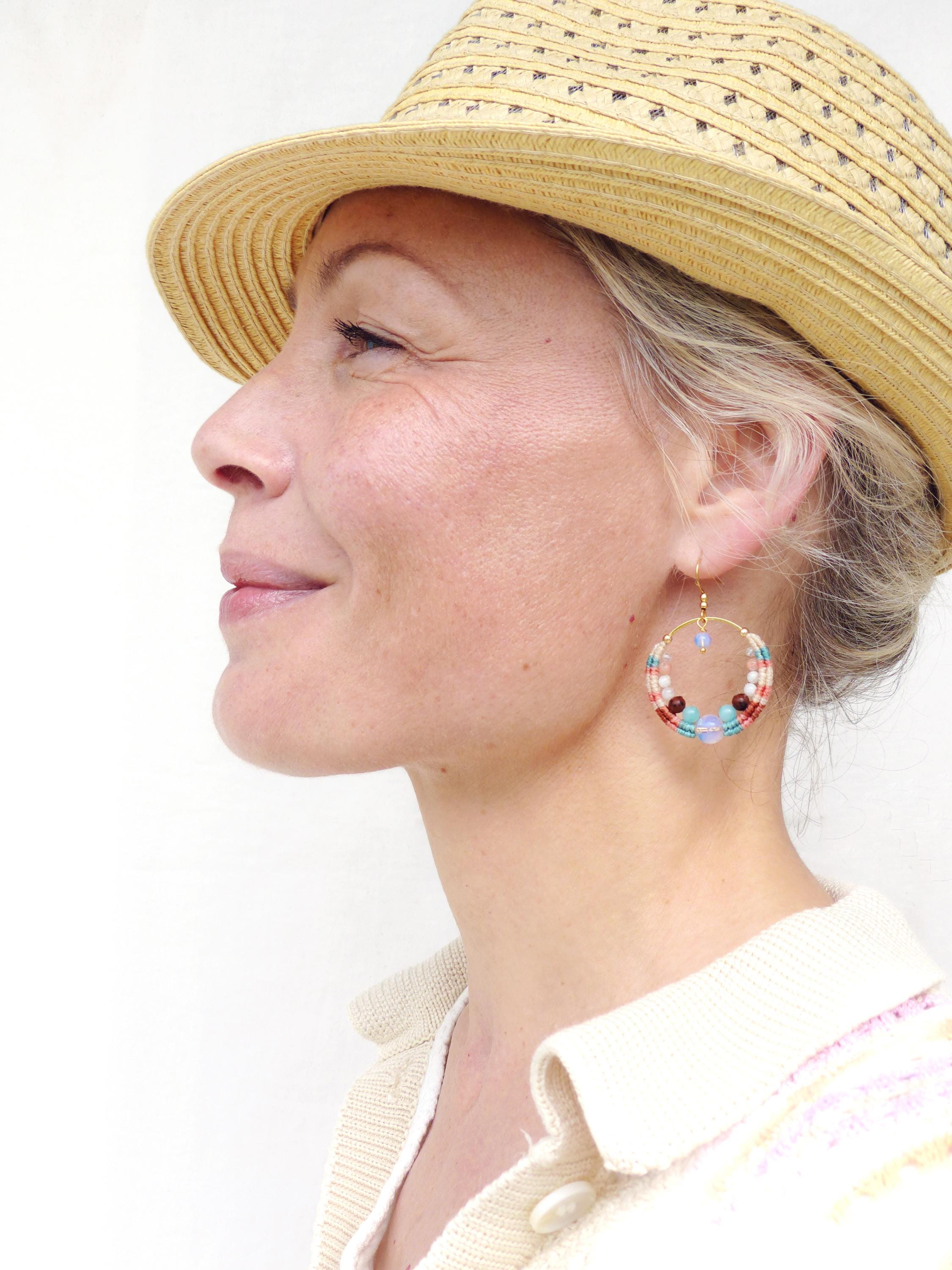 Peut inclure: Profil d'une personne portant un chapeau de paille et des boucles d'oreilles créoles dorées. Les boucles d'oreilles sont ornées de perles colorées rouges, blanches, bleues et marron. La personne porte un pull crème.