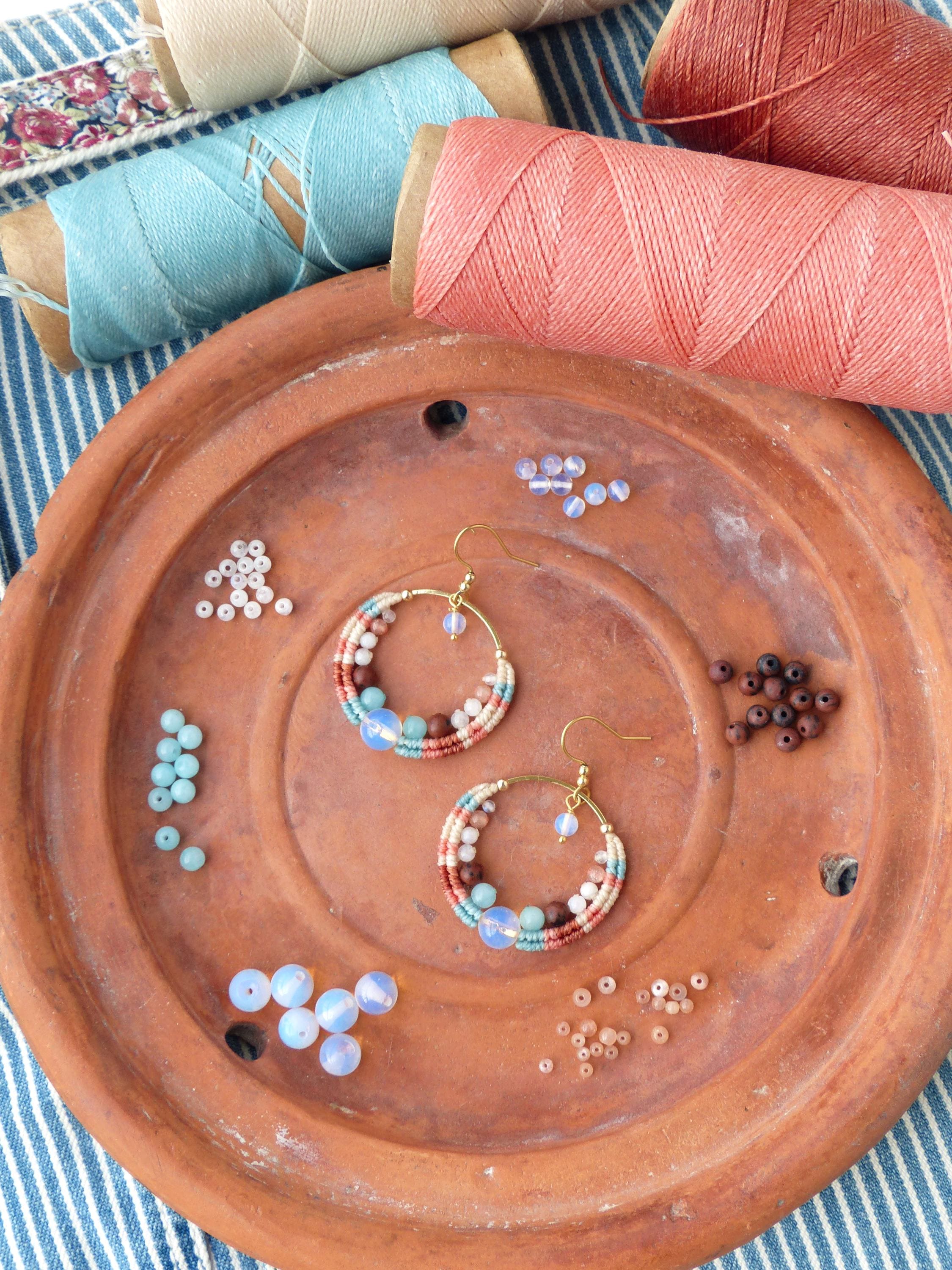 Peut inclure: Boucles d'oreilles créoles faites à la main avec un motif tissé dans des tons de rose, blanc et sarcelle, rehaussées de perles bleues et marron. Les boucles d'oreilles sont présentées sur une assiette en terre cuite avec des bobines de fil et des perles.