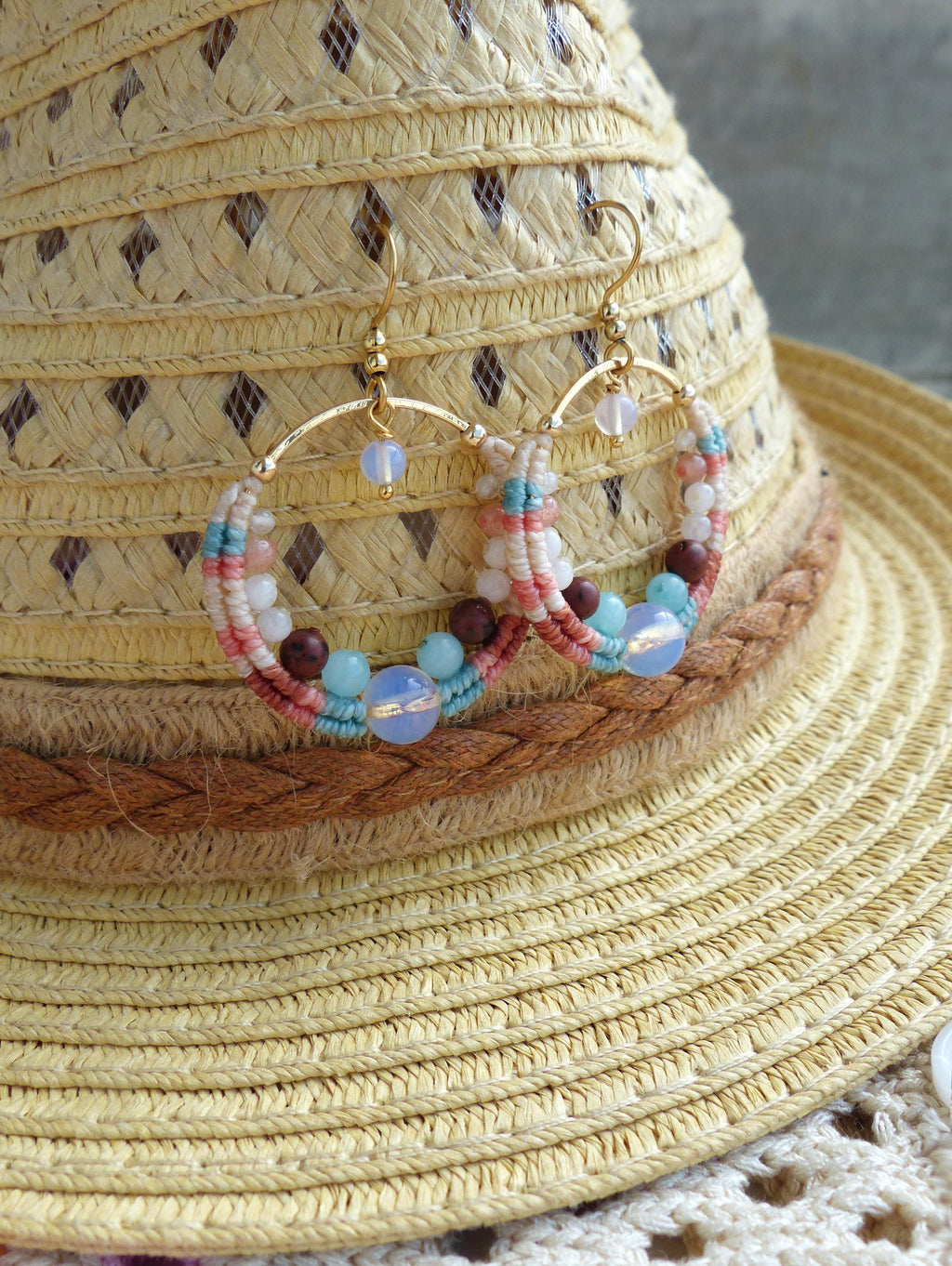 Peut inclure: Boucles d'oreilles créoles faites à la main avec un design macramé. Les boucles d'oreilles présentent un cerceau en métal doré, orné de fil blanc, rose et turquoise, et agrémenté de perles bleues, marron et blanches. Les boucles d'oreilles sont présentées sur un chapeau de paille.