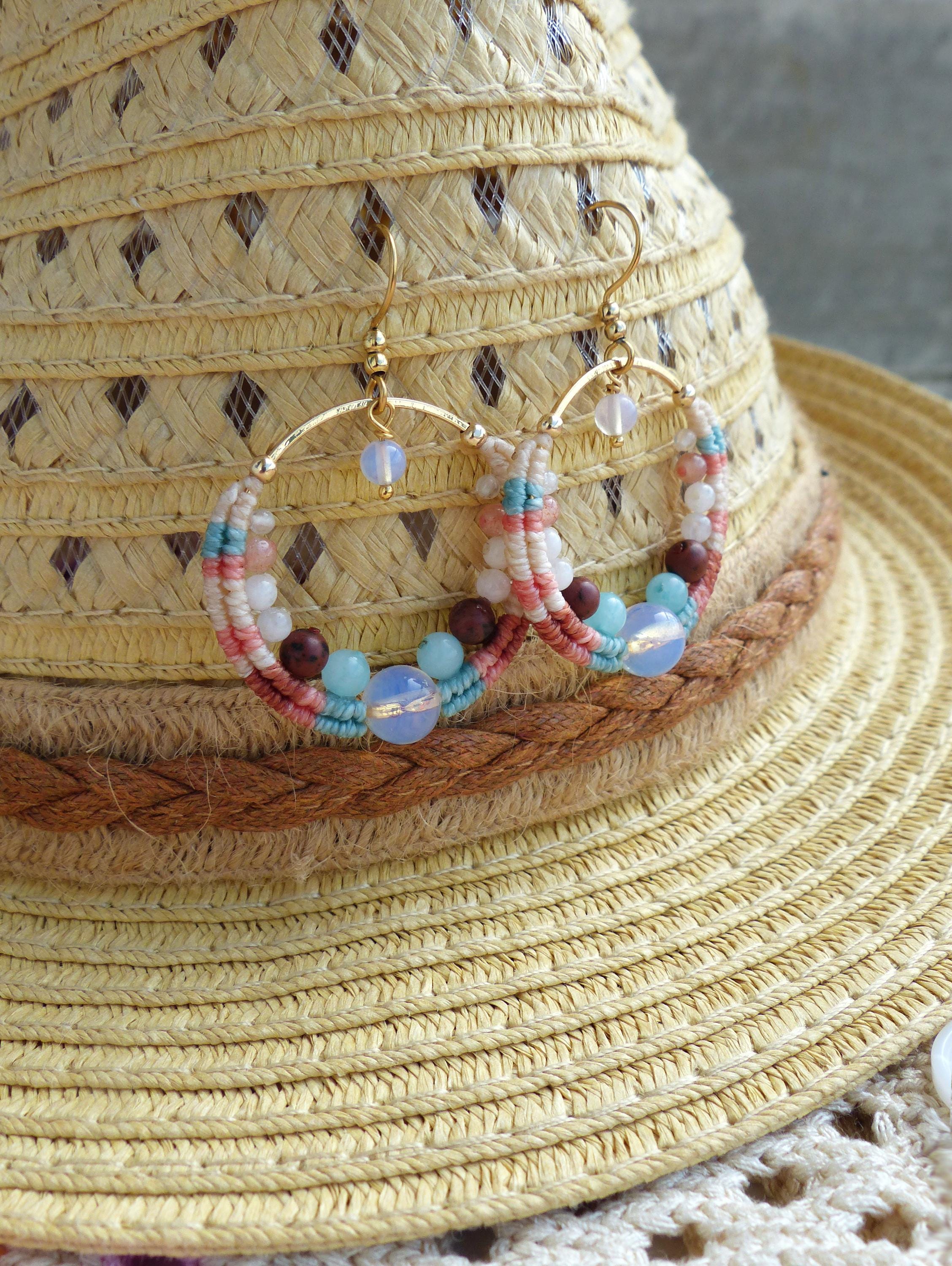Peut inclure: Boucles d'oreilles créoles faites à la main avec un design macramé. Les boucles d'oreilles présentent un cerceau en métal doré, orné de fil blanc, rose et turquoise, et agrémenté de perles bleues, marron et blanches. Les boucles d'oreilles sont présentées sur un chapeau de paille.