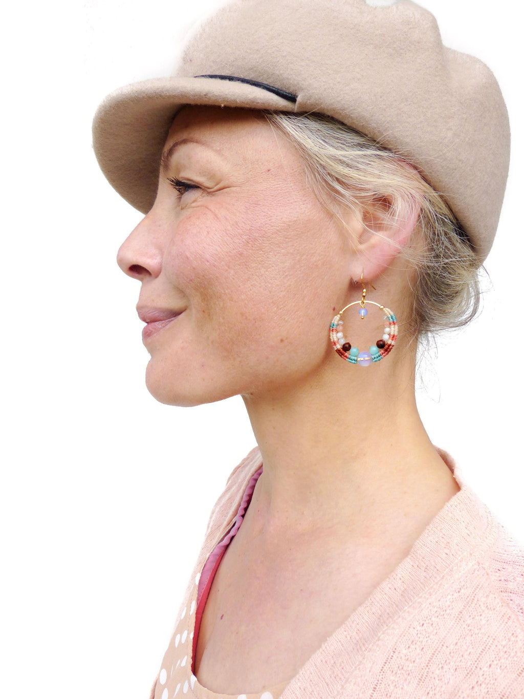 Peut inclure: Une femme porte un chapeau beige et un cardigan rose clair. Elle porte des boucles d'oreilles dorées avec des perles colorées dans les tons de rouge, bleu et blanc. Les boucles d'oreilles sont au centre de l'image.