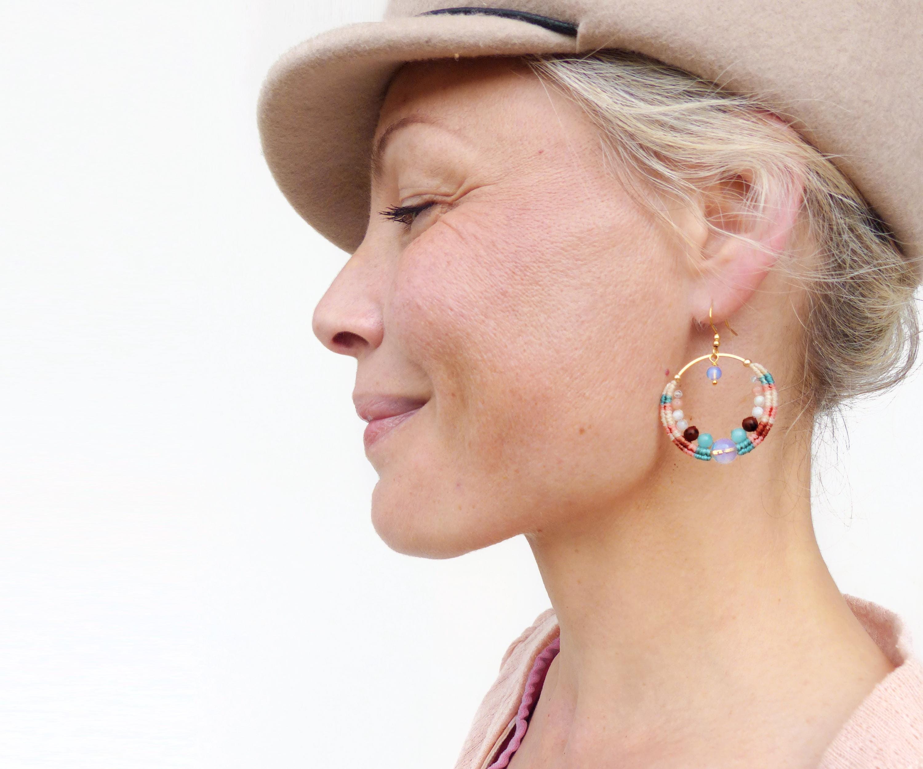 Peut inclure: Une femme porte des boucles d'oreilles créoles dorées avec des perles colorées. Les boucles d'oreilles présentent un motif circulaire avec des perles rouges, bleues et blanches. La femme porte un chapeau beige et un haut rose, sur fond blanc.