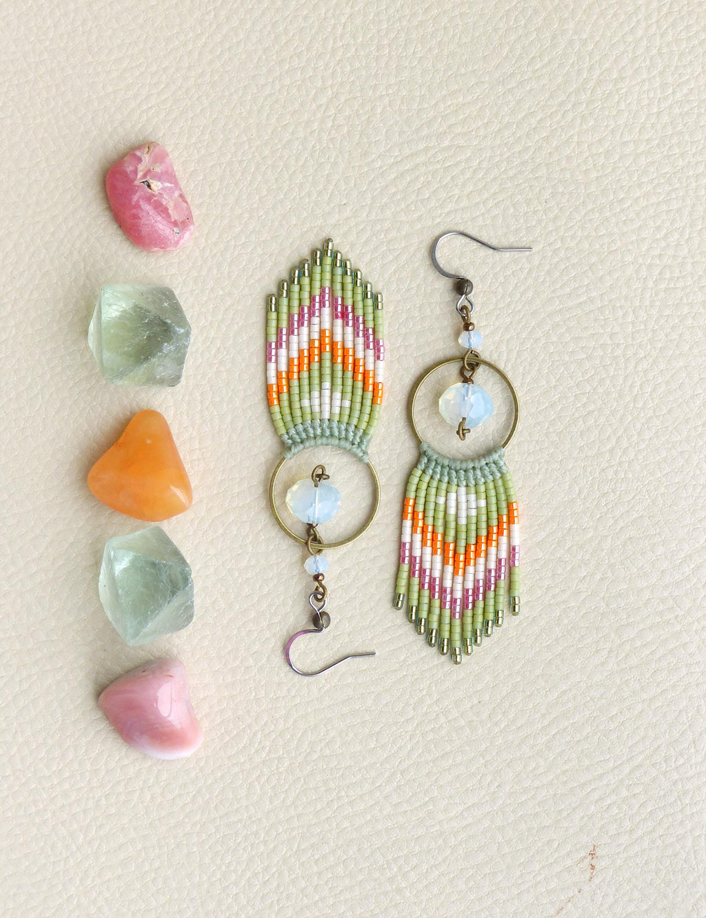 Peut inclure: Boucles d'oreilles faites à la main avec un motif en chevrons vert, blanc, orange et rose. Les boucles d'oreilles ont un design à franges, des perles transparentes et des cerceaux en métal doré. Des pierres précieuses colorées sont disposées à gauche.