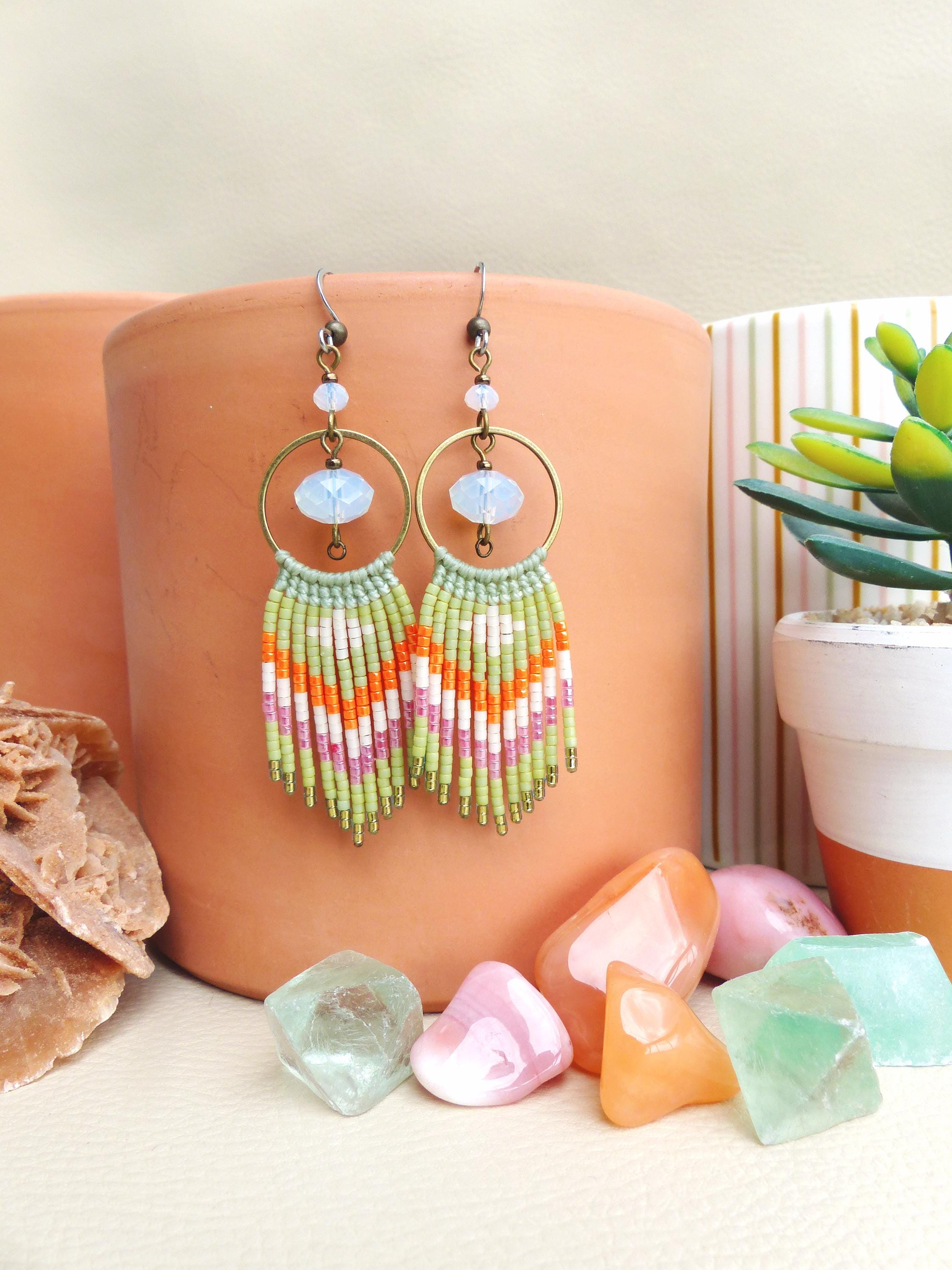 Peut inclure: Boucles d'oreilles macramé faites à la main de style bohème. Les boucles d'oreilles présentent un cercle doré, une perle bleu clair et une frange de perles vertes, oranges, roses et blanches. Elles sont présentées avec des pierres précieuses colorées et une petite plante en pot.