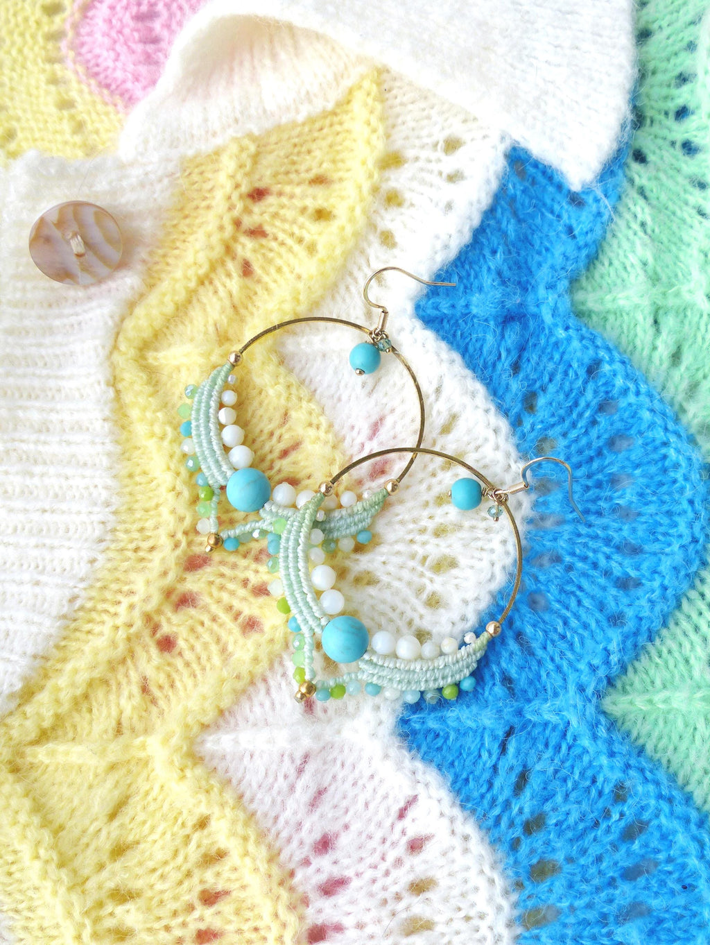 Peut inclure: Boucles d'oreilles créoles dorées ornées de perles turquoises et blanches, et d'un motif macramé vert clair. Les boucles d'oreilles sont présentées sur un tissu tricoté coloré à motifs jaunes, blancs, bleus et verts.