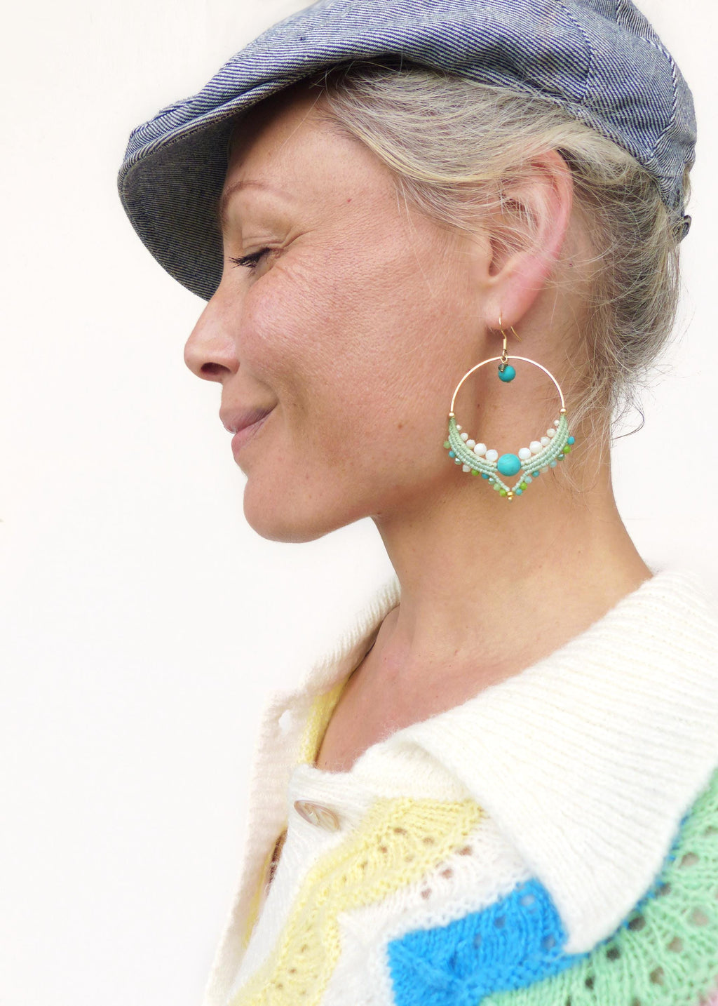 Peut inclure: Boucles d'oreilles créoles dorées avec des perles turquoise et blanches, et des détails en macramé vert. Les boucles d'oreilles sont un accessoire unique et fait à la main. La femme porte une casquette en jean bleu et un pull coloré.