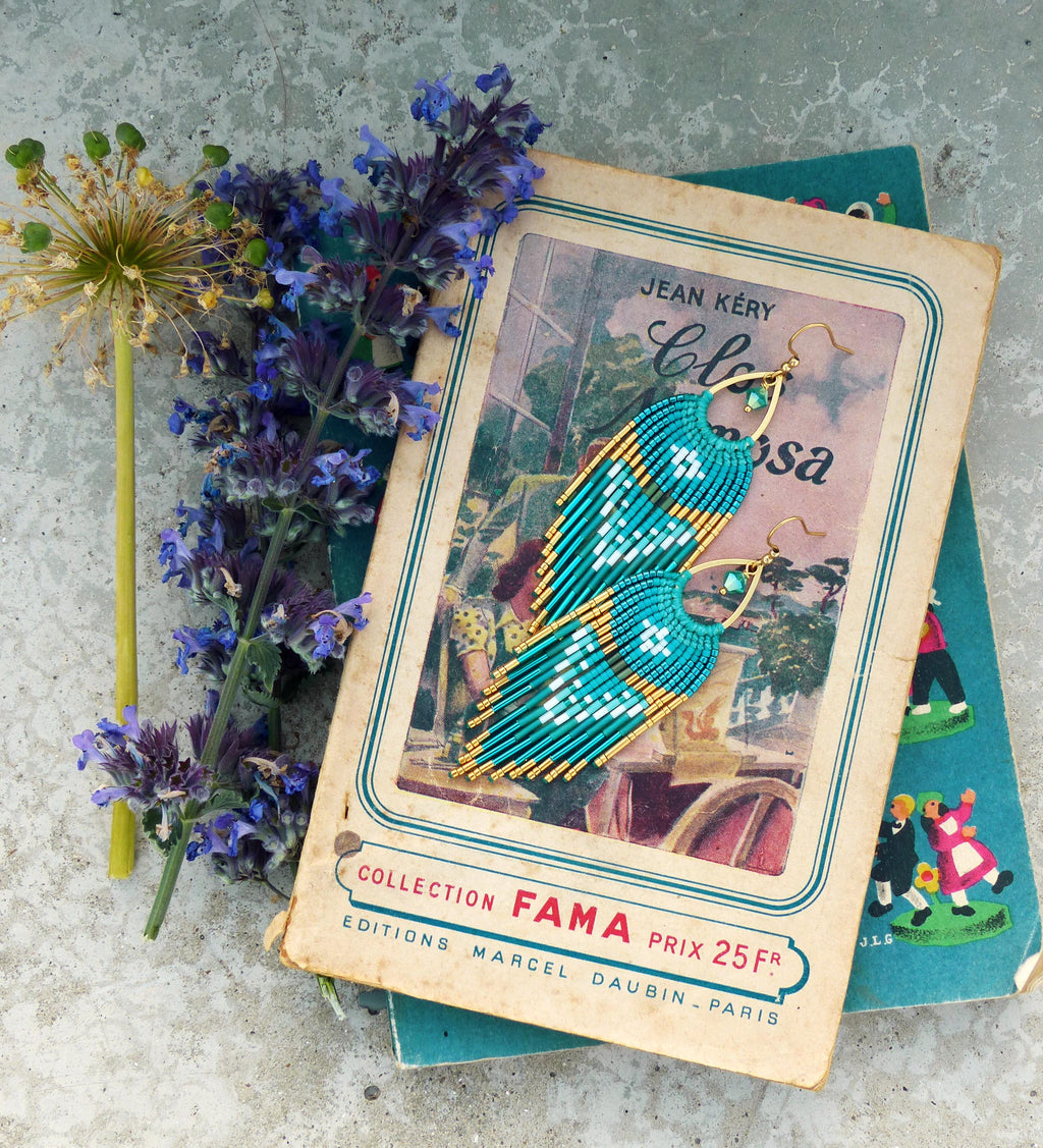 Peut inclure: Boucles d'oreilles en perles turquoise et dorées avec un motif de franges, posées sur un livre ancien. Les boucles d'oreilles présentent un motif géométrique et sont présentées avec des fleurs violettes et un livre avec du texte français.