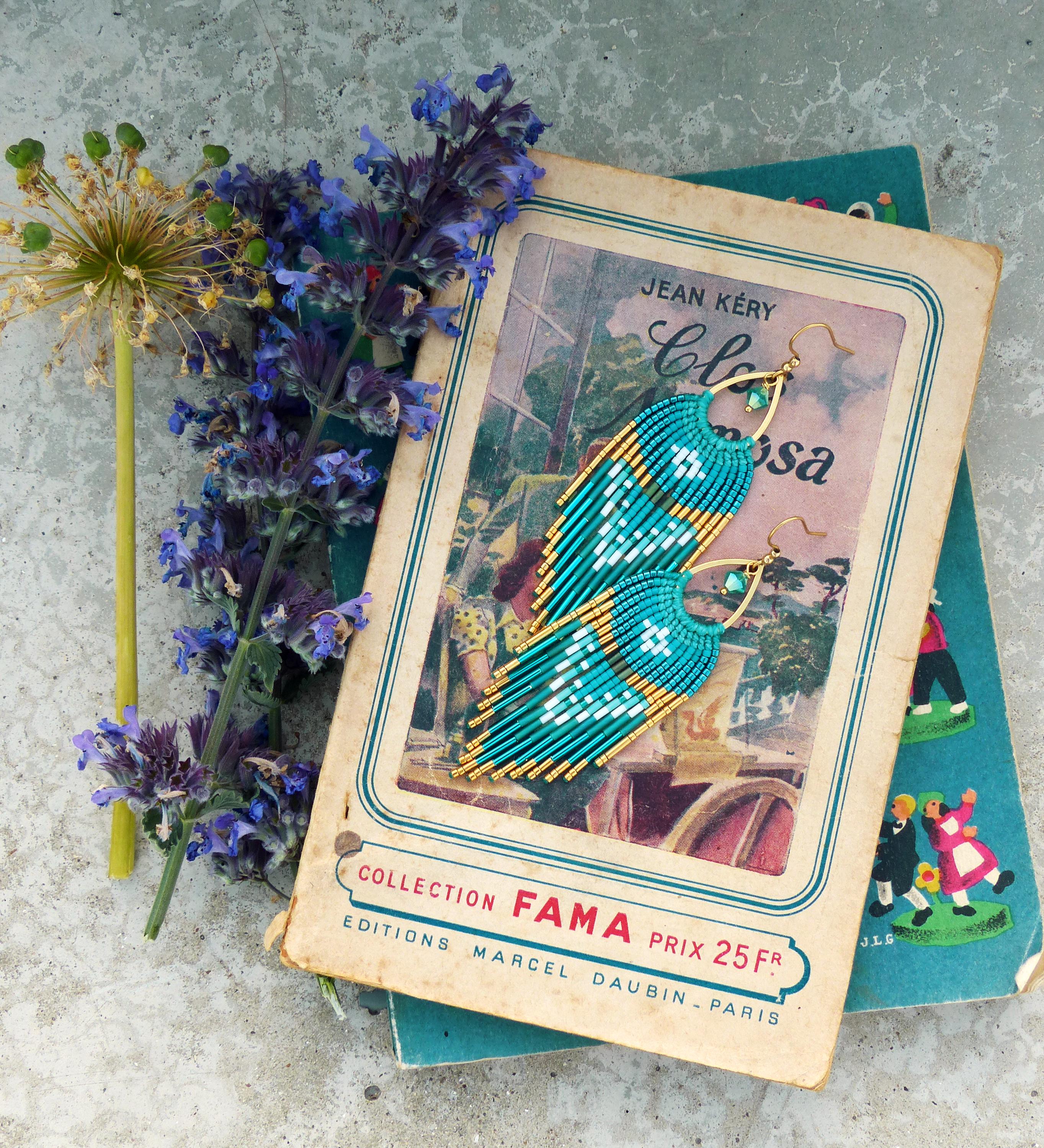 Peut inclure: Boucles d'oreilles en perles turquoise et dorées avec un motif de franges, posées sur un livre ancien. Les boucles d'oreilles présentent un motif géométrique et sont présentées avec des fleurs violettes et un livre avec du texte français.
