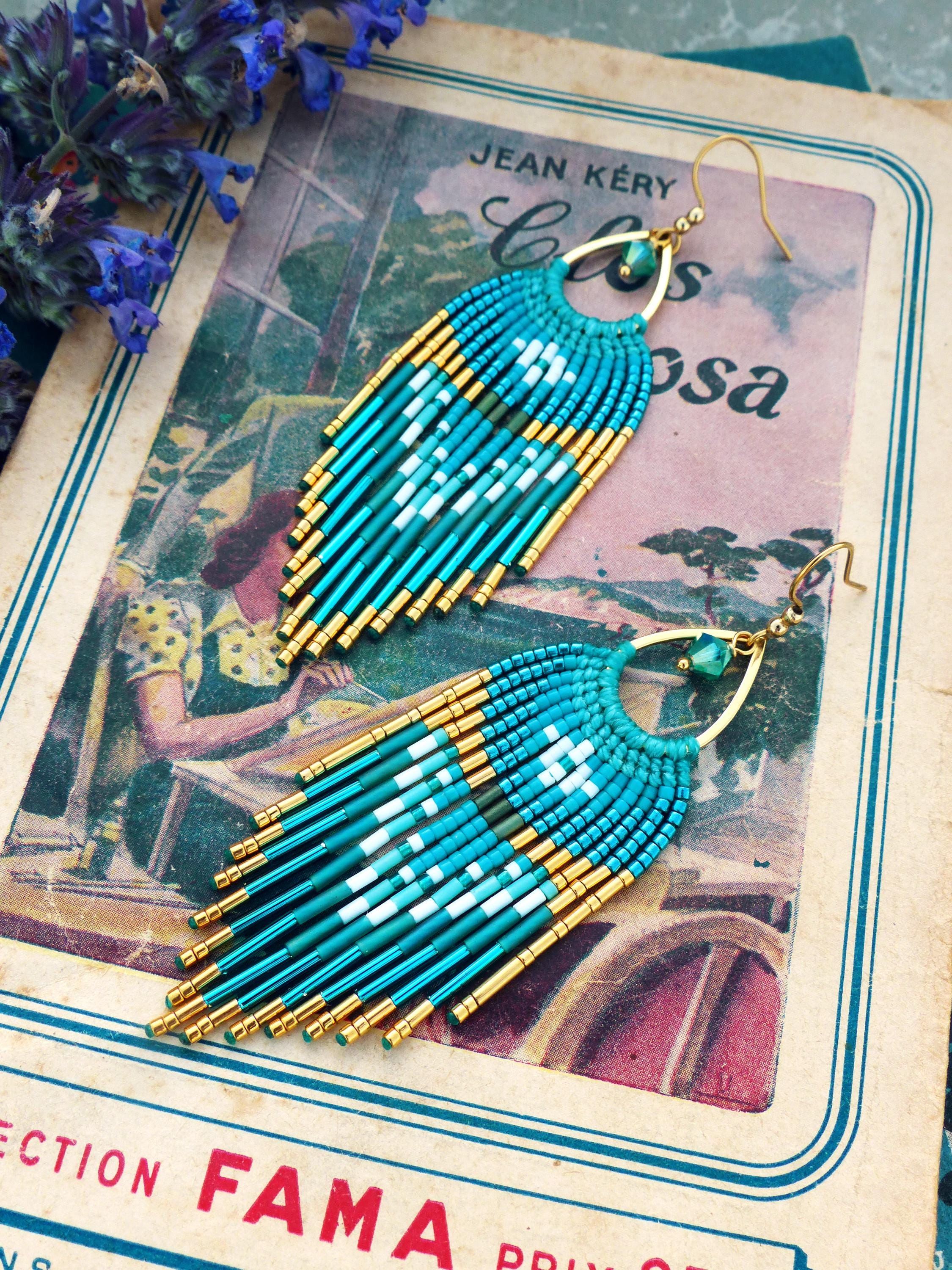 Peut inclure: Boucles d'oreilles turquoise ornées de perles dorées. Elles présentent un motif à franges avec un motif géométrique blanc et vert foncé. Elles sont présentées sur un livre ancien avec le texte "Jean Kéry" et "Collection Fama".