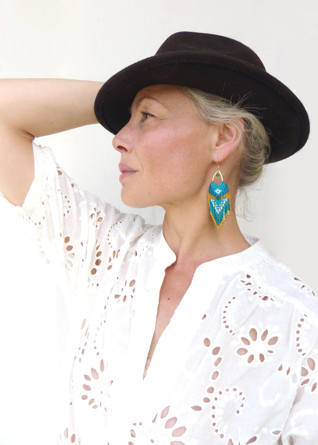 Peut inclure: Boucles d'oreilles dorées avec un motif de perles turquoise, jaunes et blanches. Les boucles d'oreilles ont une frange en bas. La personne sur l'image porte une chemise ajourée blanche et un chapeau marron foncé.
