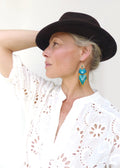 Peut inclure: Boucles d'oreilles dorées avec un motif de perles turquoise, jaunes et blanches. Les boucles d'oreilles ont une frange en bas. La personne sur l'image porte une chemise ajourée blanche et un chapeau marron foncé.
