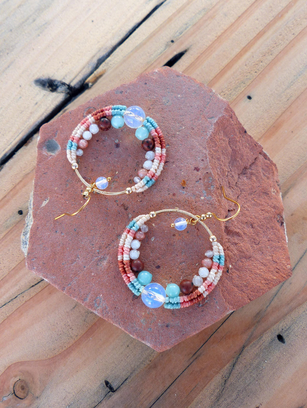 Peut inclure: Une paire de boucles d'oreilles créoles faites à la main avec un design macramé. Les boucles d'oreilles présentent un cerceau doré, enveloppé de cordon macramé rose, blanc et bleu. Elles sont ornées de diverses perles dans les tons marron, bleu et blanc. Les boucles d'oreilles sont présentées sur une pierre brun-rougeâtre.