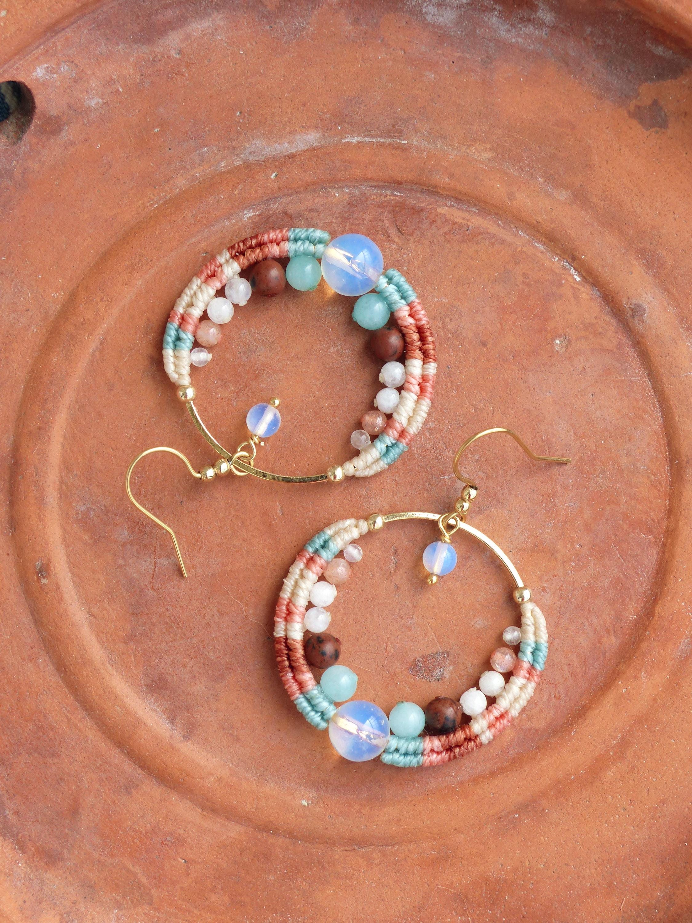 Peut inclure: Une paire de boucles d'oreilles créoles faites à la main. Les boucles d'oreilles sont composées d'un cercle en métal doré, orné de fil tissé crème, corail et turquoise. Elles sont ornées de perles blanches, marron et bleues, et d'une grosse perle opalescente.