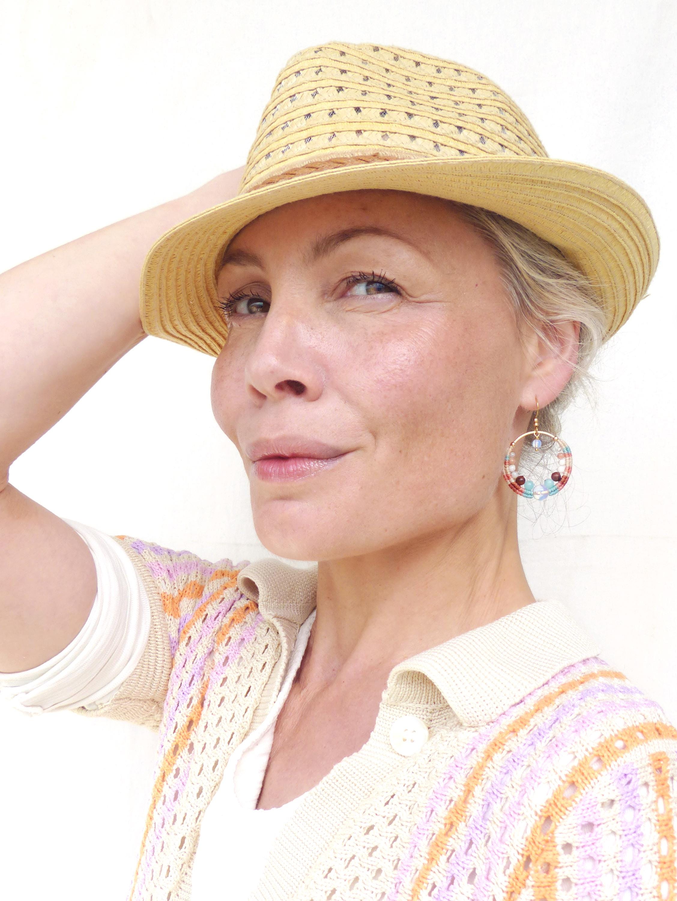 Peut inclure: Une femme porte un chapeau fedora en paille et un cardigan crème avec un motif pastel. Elle porte également des boucles d'oreilles créoles avec des perles colorées. Le fond est un mur blanc uni.