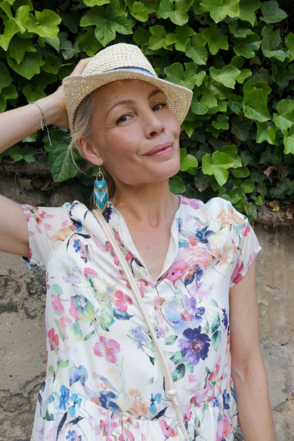 Peut inclure: Une femme porte un chapeau de paille avec un ruban bleu et une robe à imprimé floral. La robe a un col en V et présente une variété de fleurs colorées. Elle porte également des boucles d'oreilles turquoise et dorées.