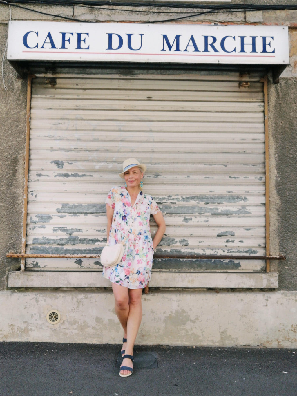 Peut inclure: Une personne porte une robe à imprimé floral, un chapeau de paille et des sandales bleues devant un bâtiment avec une enseigne "CAFE DU MARCHE". Elle porte un sac à main blanc circulaire. La robe présente un motif floral coloré.