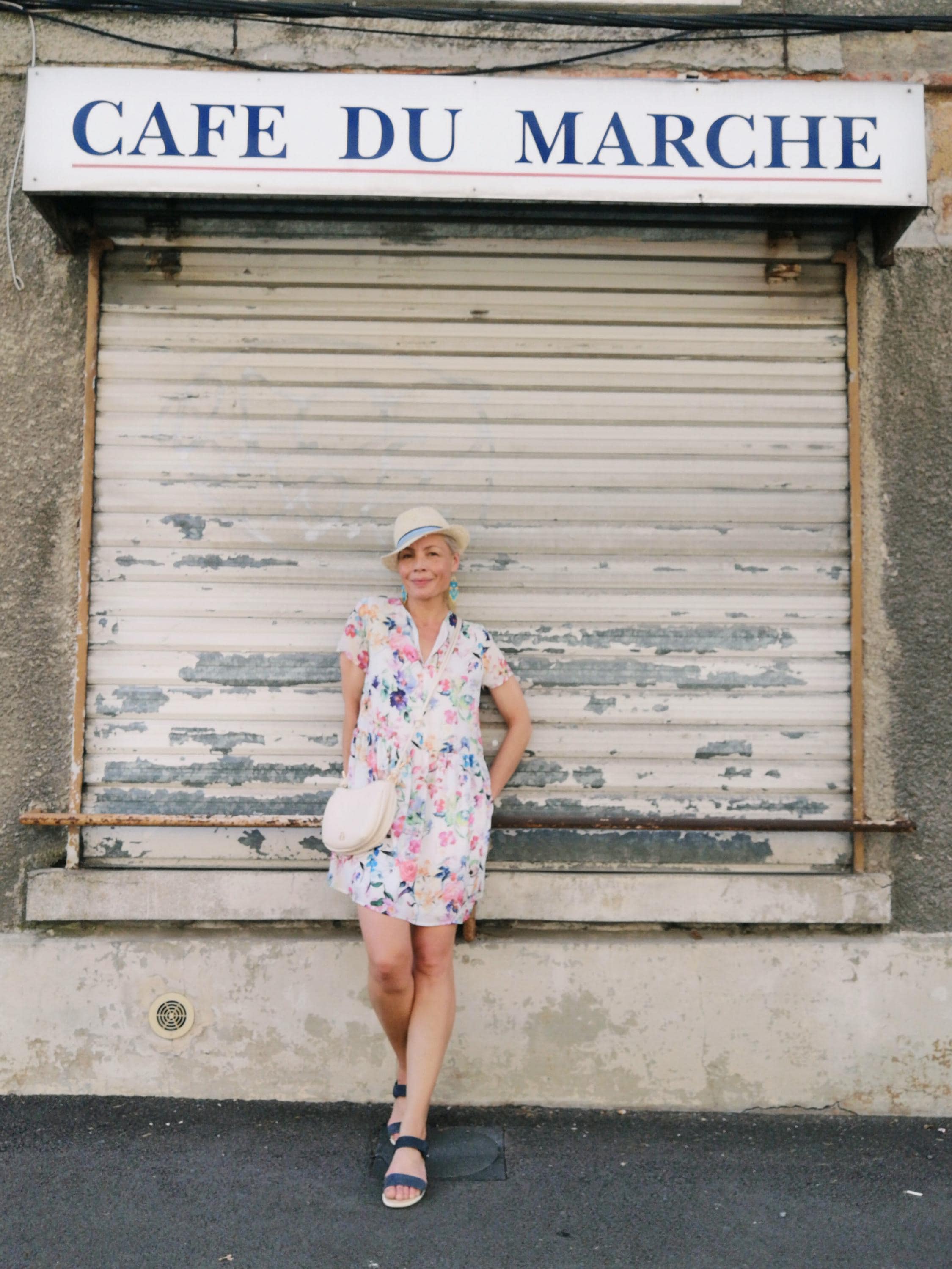 Peut inclure: Une personne porte une robe à imprimé floral, un chapeau de paille et des sandales bleues devant un bâtiment avec une enseigne "CAFE DU MARCHE". Elle porte un sac à main blanc circulaire. La robe présente un motif floral coloré.