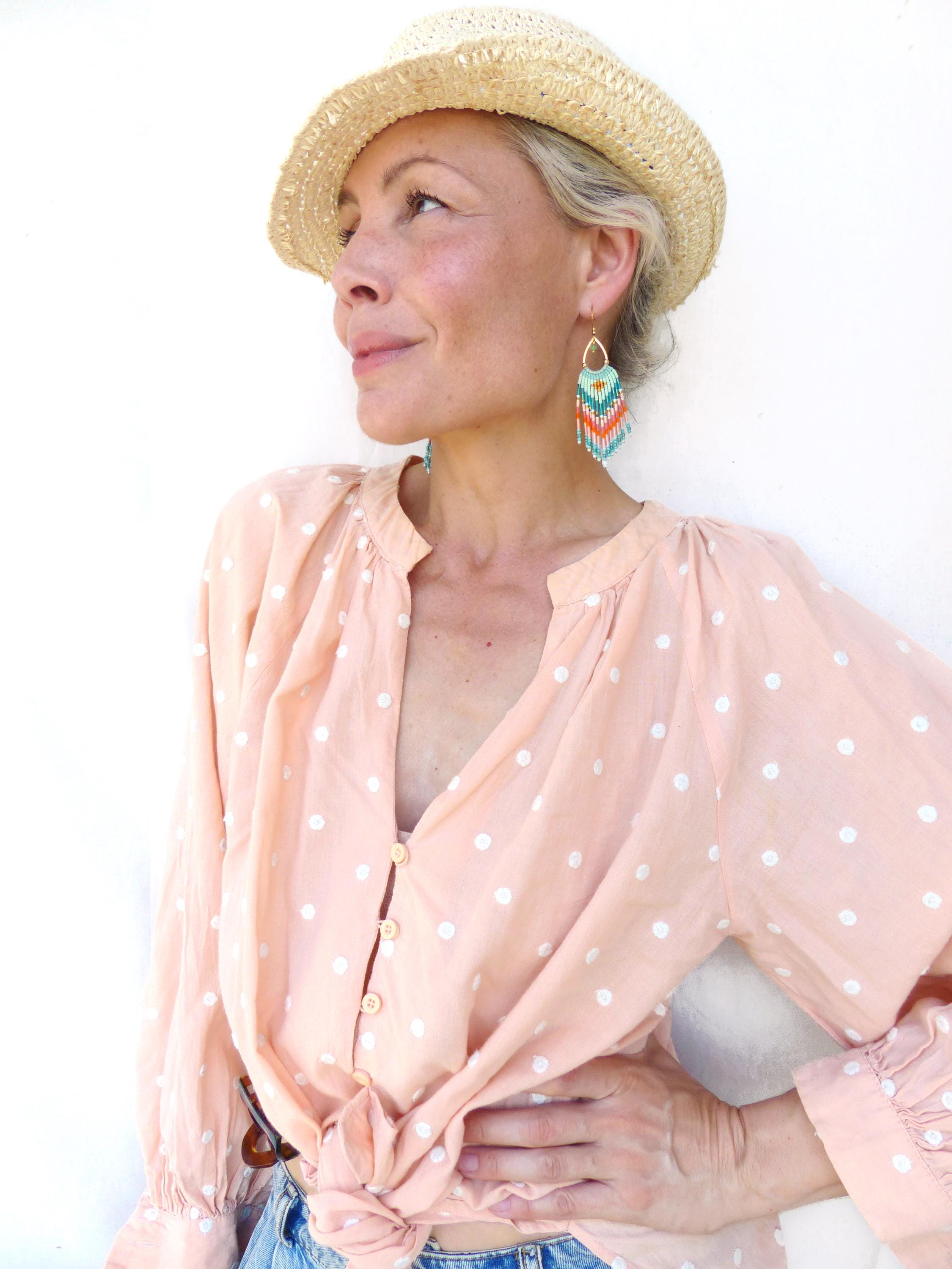Peut inclure: Une chemise boutonnée pêche clair à pois blancs, dotée de manches longues et nouée sur le devant. La chemise est associée à un chapeau de paille tressé et à des boucles d'oreilles pendantes en perles dans des tons turquoise, orange et blanc.