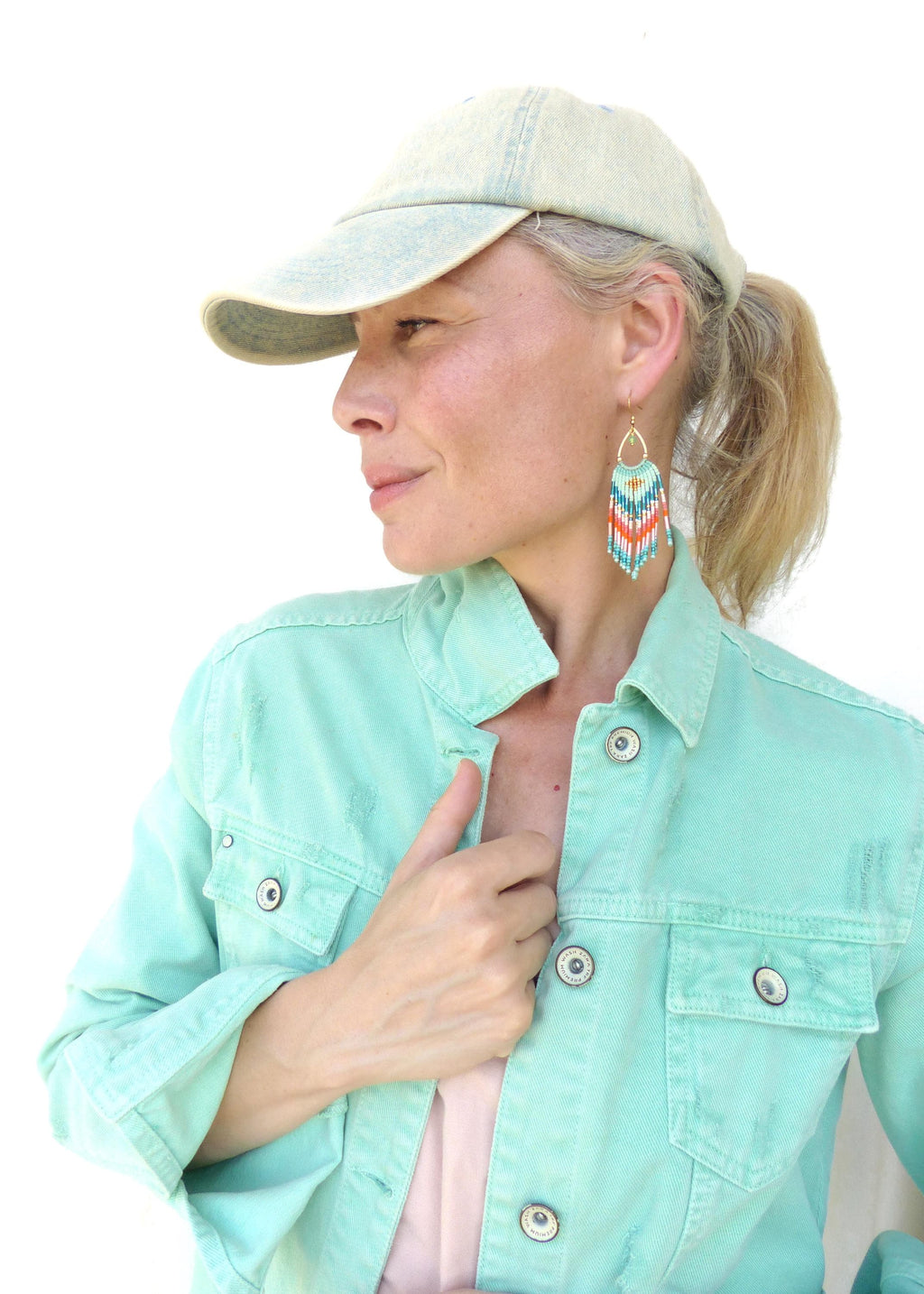 Peut inclure: Une veste en jean vert menthe clair avec des boutons argentés et une casquette de baseball en jean délavé. La boucle d'oreille est un long motif à franges en perles triangulaires avec des rayures turquoise, orange, blanches et bleu foncé.