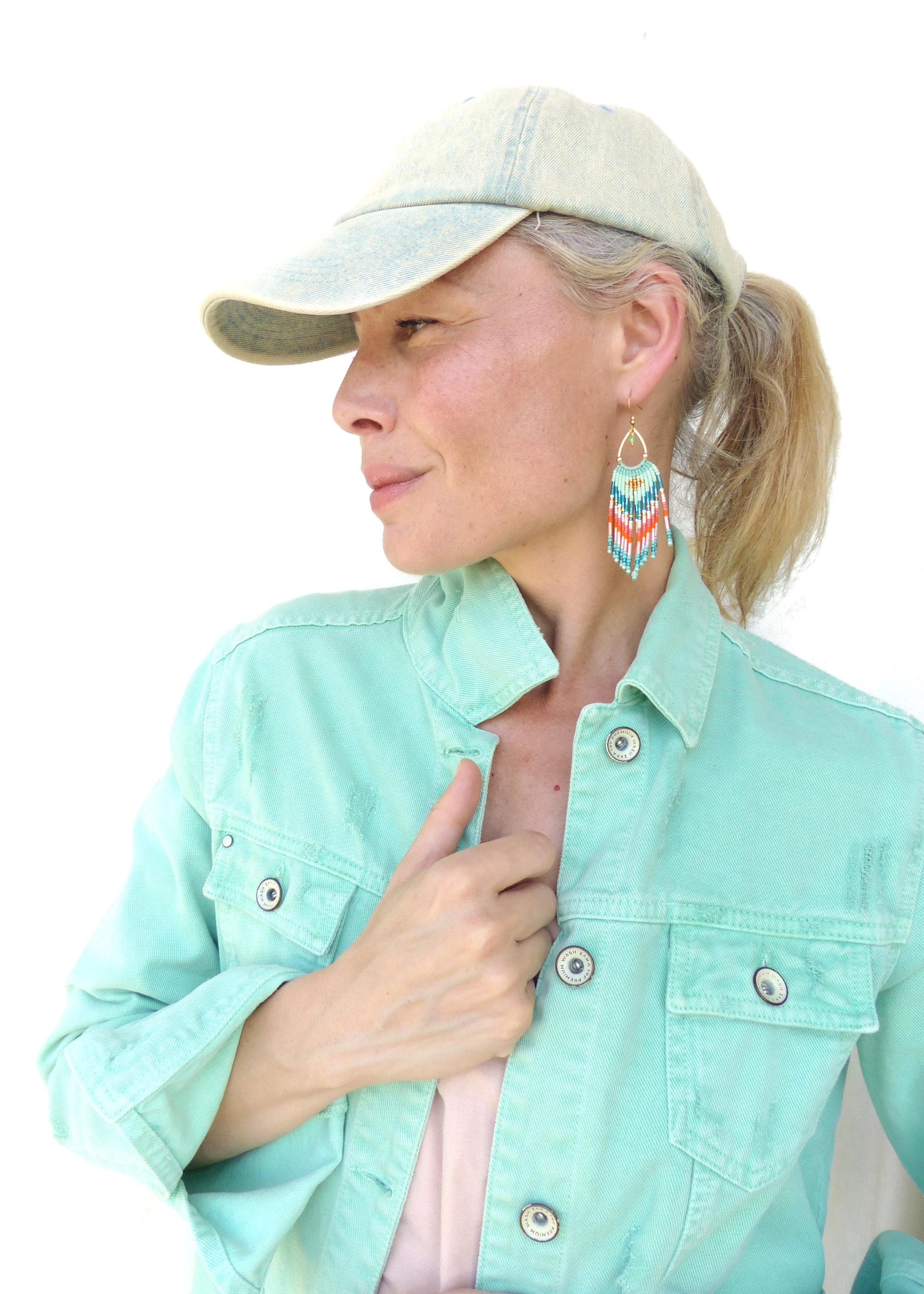 Peut inclure: Une veste en jean vert menthe clair avec des boutons argentés et une casquette de baseball en jean délavé. La boucle d'oreille est un long motif à franges en perles triangulaires avec des rayures turquoise, orange, blanches et bleu foncé.