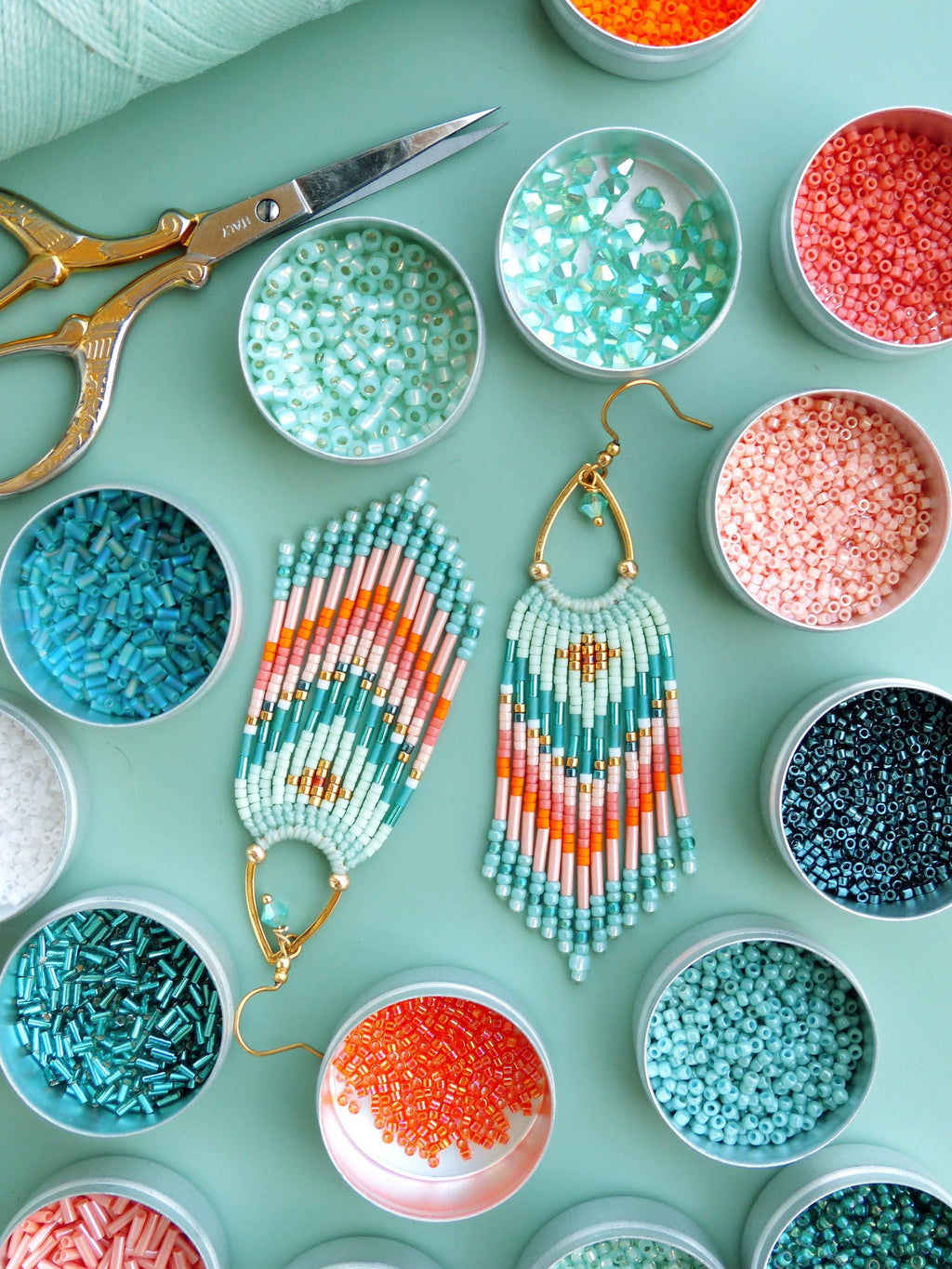 Peut inclure: Une paire de boucles d'oreilles à franges perlées présentant un motif géométrique dans des tons vert menthe, corail et doré, suspendues à des crochets dorés. Elles sont entourées de petites boîtes métalliques rondes remplies de perles.